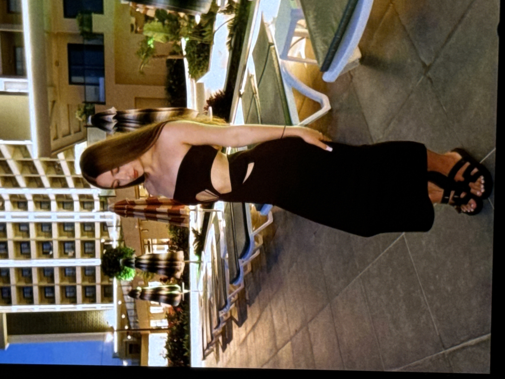 A young woman with long brown hair stands outdoors at night, posing for a photo. She is wearing a black, strapless, cut-out dress and black sandals. The background features a multi-story building, lounge chairs, and umbrellas. The scene appears to be a hotel or resort setting. There is no indication of the location being the city of Mykolaiv, Ukraine.