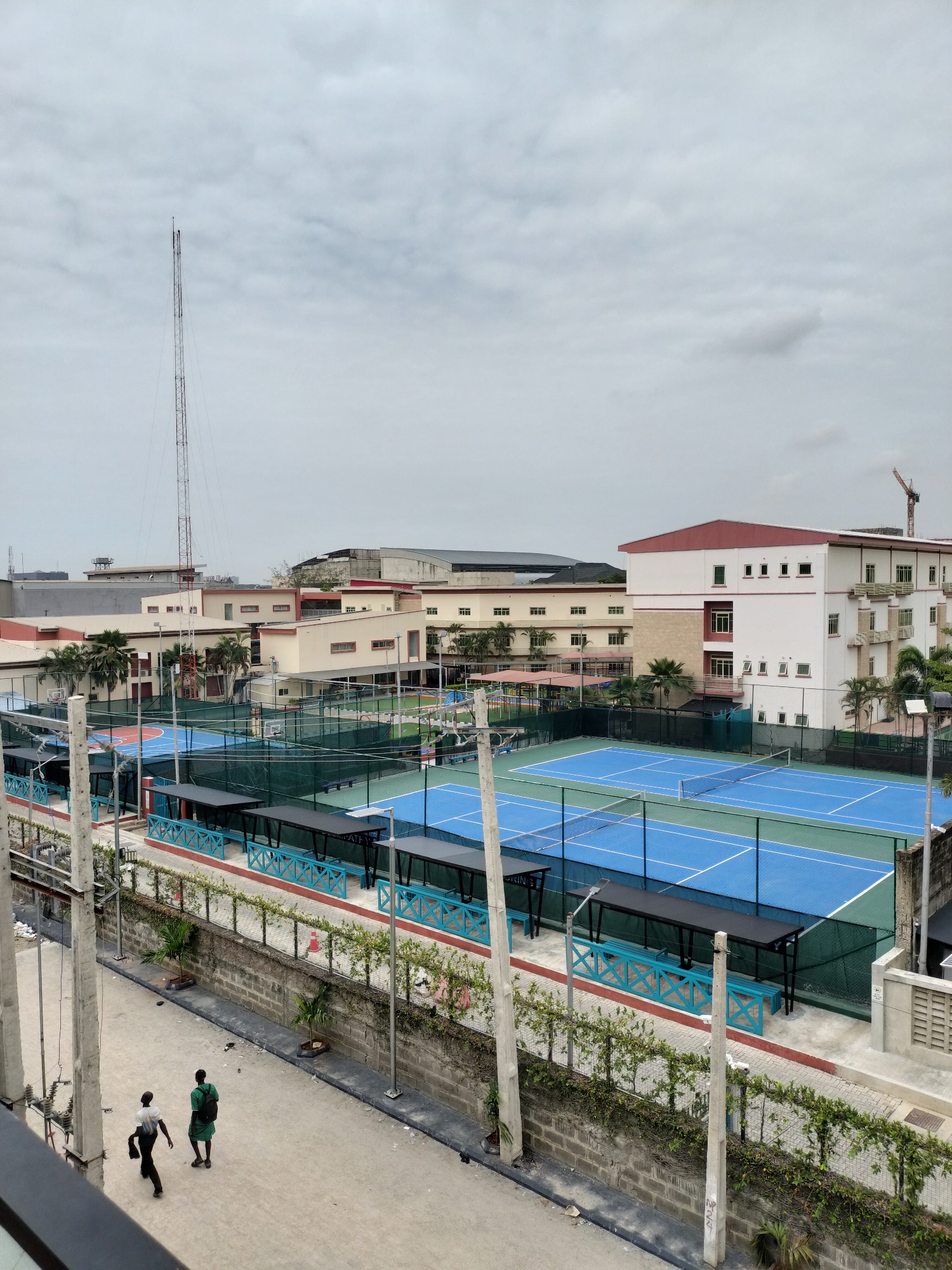 This high-angle photograph shows a section of Moba, Nigeria, featuring a complex of tennis courts and related structures. The courts are painted in blue and green, with green netting surrounding each one. Several covered benches with turquoise frames are positioned along the sides of the courts.
Beyond the tennis complex, there is a cluster of buildings with light beige exteriors and red roofing. A tall communication tower stands on the left side of the frame, and a crane is visible in the distance on the right. The sky is overcast with dense cloud cover.
In the foreground, a paved walkway shows two men, one wearing a dark shirt and trousers and the other wearing a green shirt and dark shorts, walking away from the viewer. The walkway is bordered by a low brick wall, with climbing plants and small trees along its length. Concrete poles with electrical wiring are also present along the side of the walkway.