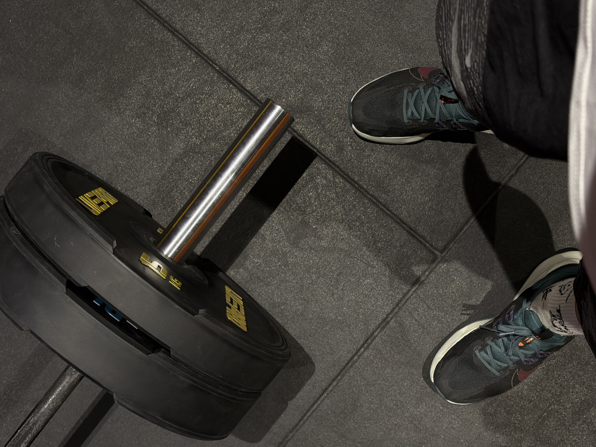 A top-down view shows a barbell with black weight plates, next to a person wearing dark athletic shoes. The barbell is partially visible on the left, loaded with at least two black weight plates with gold lettering. The person is standing to the right of the barbell, wearing running shoes, dark workout pants, and visible white socks. The scene is set on a dark gray tiled floor, likely inside a gym.
I cannot verify a specific location in Krasnoyarsk, Russia. The image is a general fitness scene.