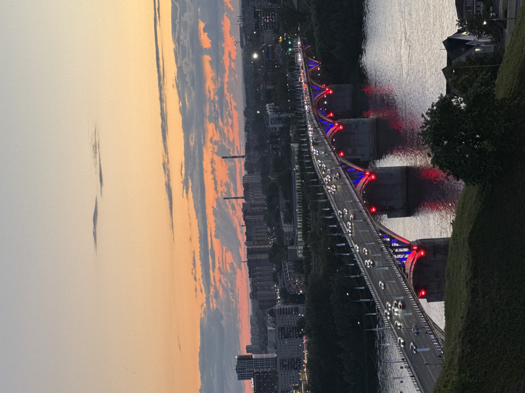 The image depicts a panoramic view of the city of Nizhny Novgorod, Russia, at dusk. A prominent, illuminated bridge spans across a body of water, with a continuous stream of car headlights and taillights visible on its roadway. The bridge is adorned with red and blue lights along its underside and supporting structures, casting a vibrant glow on the water.
In the background, the city skyline is characterized by a dense arrangement of buildings, varying in height and architectural style, with some taller, modern structures interspersed with older, lower-rise buildings. Industrial chimneys are also visible on the horizon, indicating an industrial presence within the city. The sky above is a canvas of gradient colors, transitioning from a soft, pale orange near the horizon to a muted blue or grey higher up, with scattered clouds catching the last light of the setting sun.
The foreground features a grassy embankment with some trees, suggesting an elevated vantage point from which the photograph was taken. The overall atmosphere is one of a bustling city at twilight, with the illuminated bridge serving as a central focal point. No people are clearly discernible in the image, and the primary action is the movement of vehicles across the bridge. The environment is urban, with elements of both nature (trees, water, sky) and human infrastructure (buildings, bridge, vehicles).
