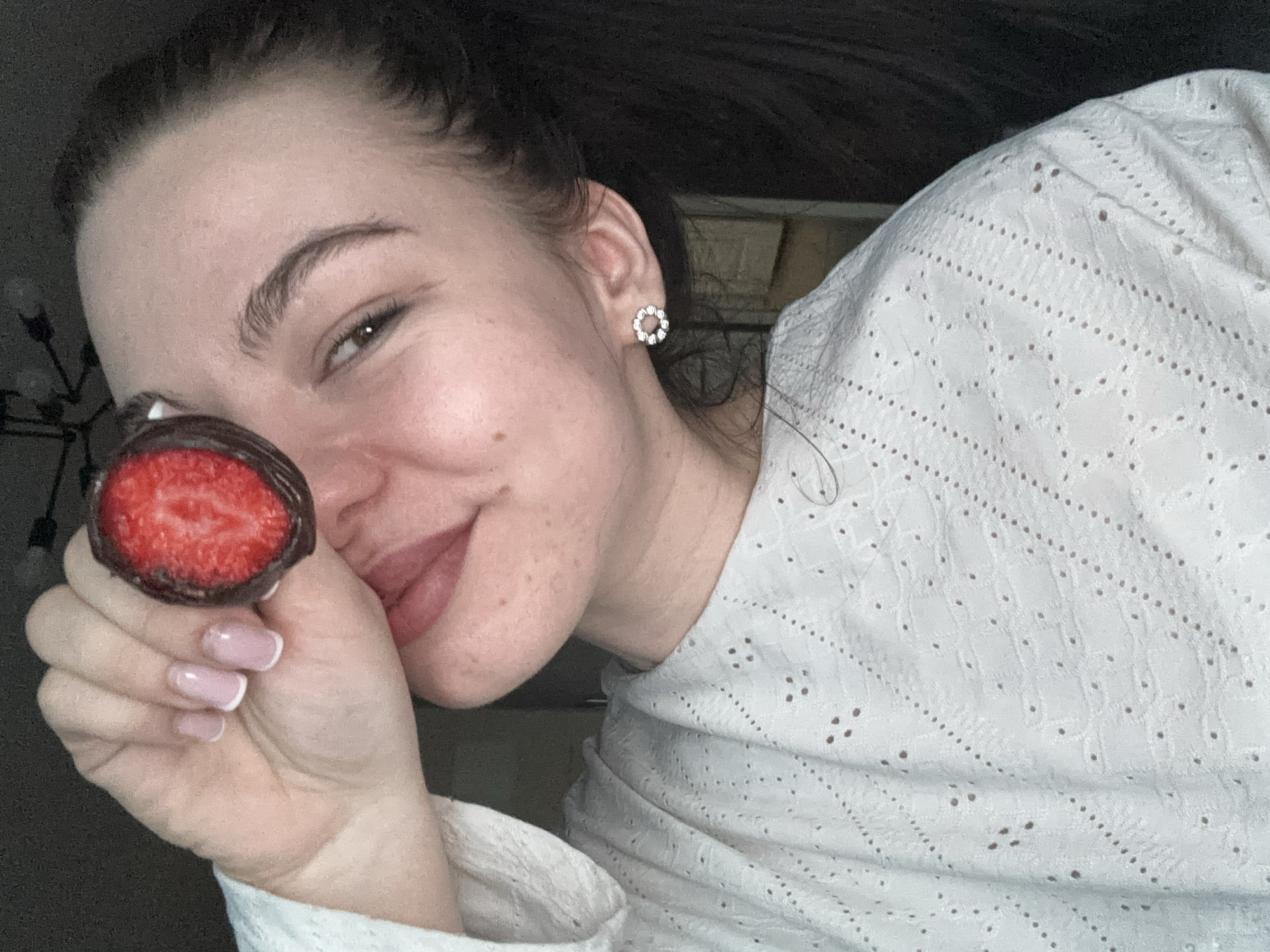 A young adult female is depicted in a close-up, indoor portrait originating from Perm, Russia. She is smiling, with her lips slightly parted, and her dark hair is pulled back into a ponytail visible on the right side of the frame. Her right eye is visible, while her left eye is obscured by a vertically halved strawberry. The strawberry is partially coated in dark chocolate on its exterior, with its exposed red, seeded interior facing the viewer. Her right hand, exhibiting French-tipped manicured nails, holds the strawberry near her face. She wears a white, eyelet-patterned top and a circular, diamante-studded earring in her right ear. The background is dark and out of focus, with a faint, dark, decorative fixture visible in the upper left quadrant.