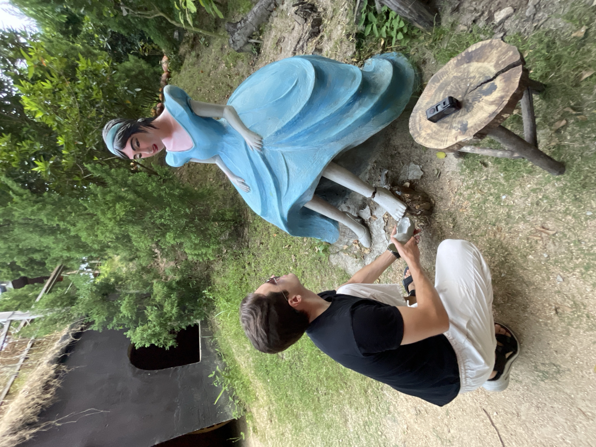 An outdoor scene, likely a themed garden or park in Nam Nha Trang, Vietnam, features a life-sized statue of a woman in a flowing light blue gown and a person interacting with it. The original image is rotated 90 degrees counter-clockwise.
The statue, possibly depicting a fairy tale character like Cinderella, has dark hair, a gentle expression, and bare feet. It appears to be leaning forward slightly, with its elaborate blue dress creating a sense of movement.
In front of and looking up at the statue is a person, likely male, wearing a black t-shirt, white pants, and black sandals. They are crouching or kneeling, holding a small, light-colored, possibly broken object in their hands, seemingly in the process of placing or presenting it to the statue's bare foot. Sunglasses are pushed up onto their head.
To the right of the statue, a small, rustic wooden table, made from a tree stump with three rough legs, holds a black action camera. The background is densely filled with lush green foliage and trees, creating a natural and verdant setting. In the lower left quadrant of the image, a dark opening, possibly an entrance to a cave or a structure, is visible. The scene is illuminated by natural daylight. No visible text is present.
