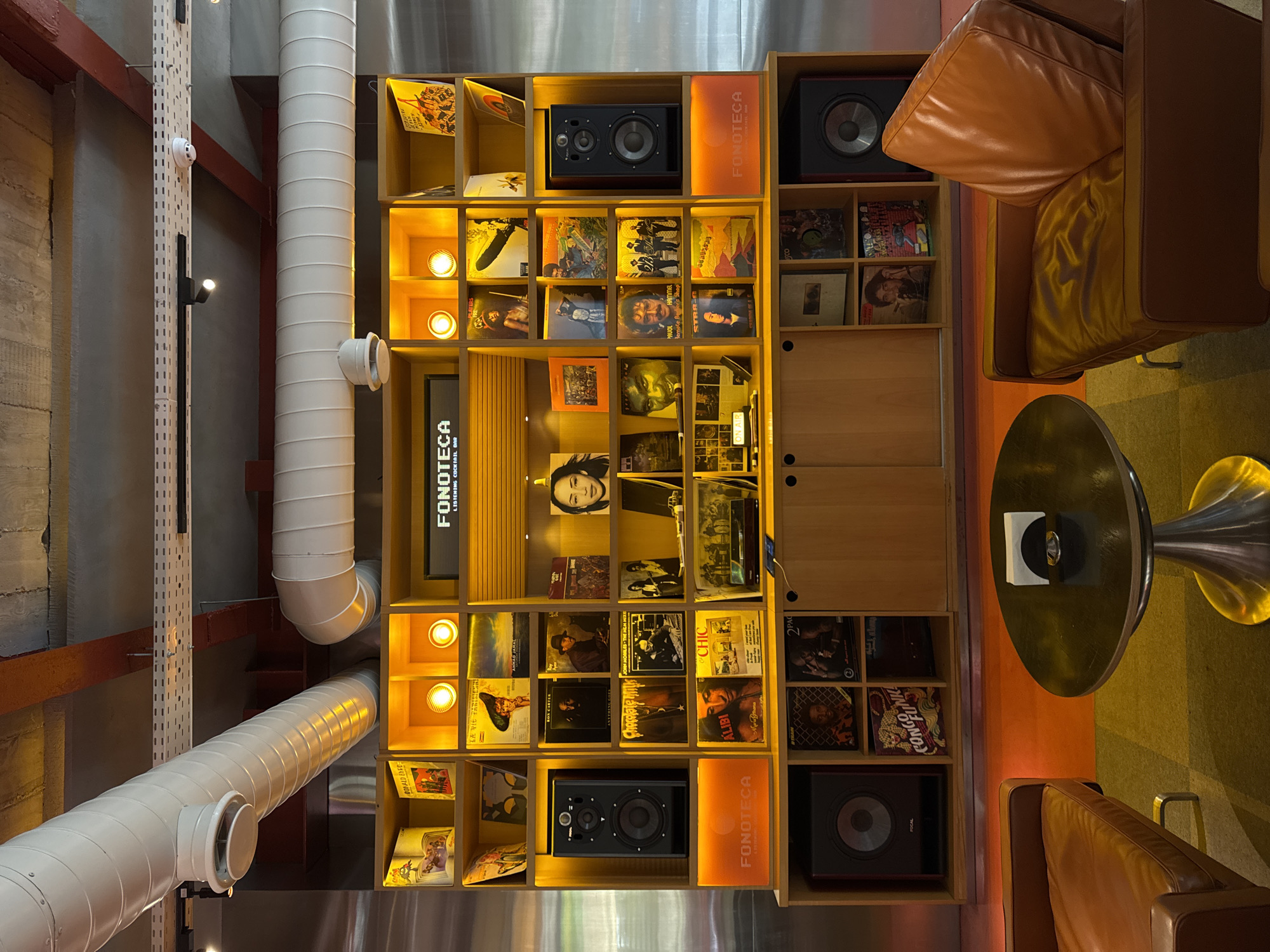 The image captures the interior of "Fonoteca," a listening cocktail bar located in Yerevan, Armenia. The scene is dominated by a large wooden shelving unit filled with vinyl records and audio equipment. Two brown leather armchairs and a metallic table with a circular top are positioned in the foreground, suggesting a comfortable seating area. The shelving unit features various compartments, some housing speakers, others displaying record album covers, and some illuminated by warm lighting. A prominent "Fonoteca" logo is visible on the unit. Above the shelves, large ventilation pipes are attached to the ceiling, contributing to the industrial aesthetic of the space. The overall impression is a stylish and retro-themed establishment focused on music and drinks.