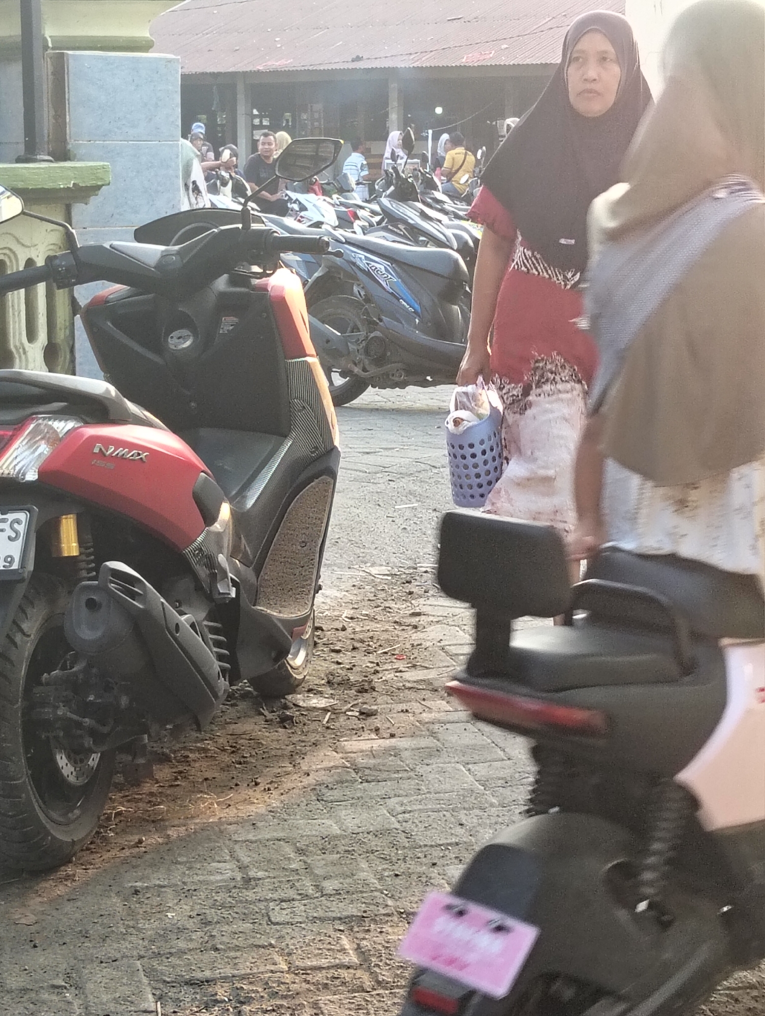 This is a medium shot, eye-level, outdoor photograph taken in Banjarwati, Indonesia, featuring several parked motorcycles and two women. The foreground is dominated by the rear and side of a red scooter, identified by the "NMAX" logo. The scooter has a black footrest with a patterned texture and a yellow shock absorber visible. To the right and slightly behind the red scooter, another scooter is partially visible, with a black seat and part of a pink license plate.

In the background, a woman wearing a black hijab and a red and white patterned dress is holding a blue woven basket filled with items. She is looking to her left. Partially obscuring her from the right is another person, whose face is not visible, wearing a gray garment. Behind them, several other motorcycles are parked, and a group of people are gathered near a building with a corrugated metal roof. The ground is paved with bricks and covered in dirt and debris. The lighting suggests it is daytime, possibly late afternoon due to the warm glow.