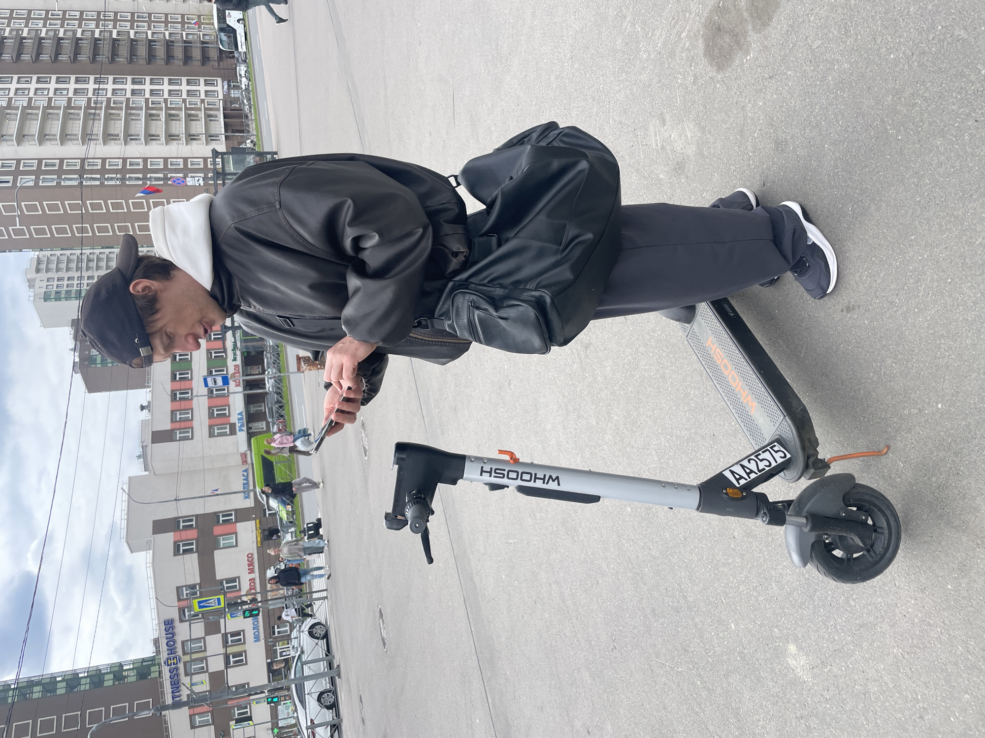 A person is standing on a scooter and looking at a mobile phone in an urban area. The individual is wearing a black jacket, dark pants, and black and white sneakers, with a black baseball cap and sunglasses. A white hoodie is visible under the jacket. They are carrying a black bag across their body. The scooter is gray and black with the word "WHOOSH" printed on it. The background includes buildings with multiple windows, street signs, traffic lights, cars, and other pedestrians. In the distant background, a building with "FITNESS HOUSE" on it is visible. The location is Unknown, Russia.