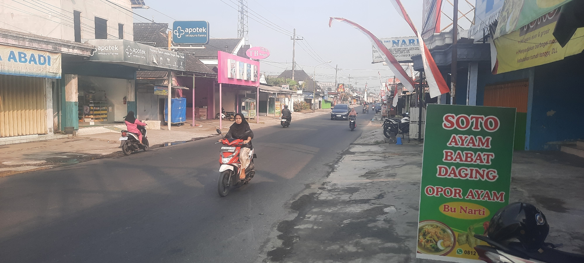 This is an eye-level, outdoor shot of a street in Selopuro, Indonesia, featuring a road with several motorcycles and a car. The buildings lining the street appear to be small shops, with one prominently displaying a sign for "Apoter Selupuro Farma," a pharmacy. Another shop has a pink facade with a sign reading "TB Denalw." A large green sign in the foreground advertises "Soto Ayam Babat Daging Opor Ayam Bu Narti," indicating a food establishment. Two women are riding motorcycles, one in the foreground on a red and white scooter and another further back on a pink scooter. The overall scene depicts a commercial street during daylight hours, with a mixture of pedestrians and vehicular traffic. The environment appears somewhat hazy, possibly due to the time of day or atmospheric conditions. Indonesian flags are visible on some of the buildings, suggesting a national holiday or celebration.