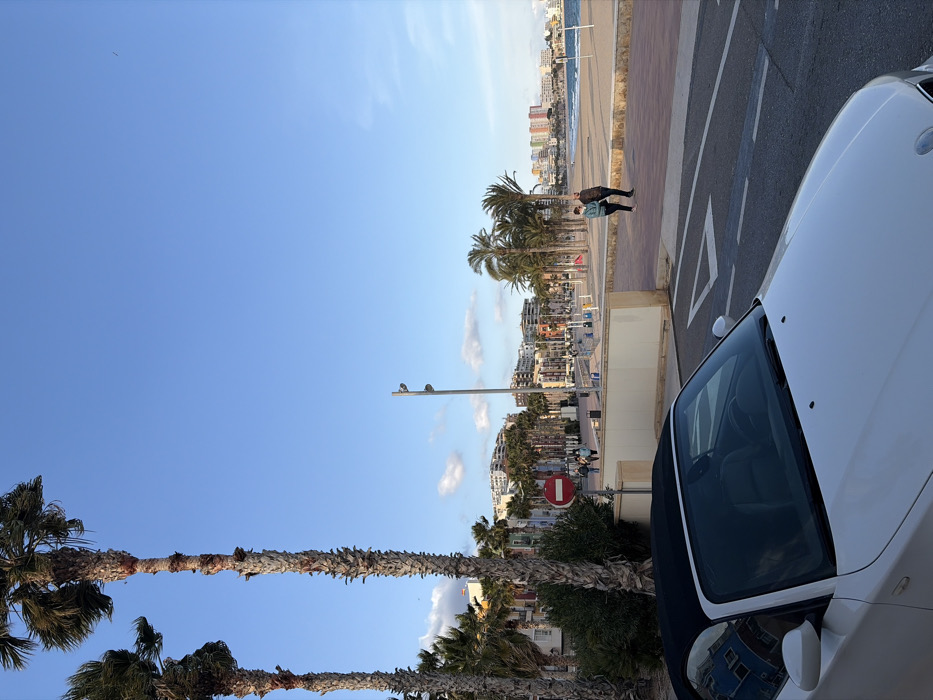 The image depicts a coastal street scene in Villajoyosa/la Vila Joiosa, Spain, under a clear blue sky with scattered white clouds. In the immediate foreground, the front hood and windshield of a white convertible car, likely a BMW Z4, with its black soft top deployed, are visible, angled slightly towards the right. White road markings, including an arrow and lane dividers, are visible on the asphalt in front of the car.
Beyond the vehicle, a beige-colored barrier separates the road from a wide promenade. Tall, slender palm trees with visible fibrous trunks line the left side of the scene and extend into the midground. Lush green foliage is present at the base of these trees. A red circular "No Entry" sign with a white horizontal bar is positioned near the left side of the promenade.
Further along the promenade, multiple streetlights are visible. In the midground, a row of multi-story buildings with varied, colorful facades (red, yellow, blue, white) lines the street. Two individuals are walking on the red-tiled promenade on the right side; one wears a teal-colored top and dark pants, the other a darker jacket and pants. Other figures are discernable walking further down the promenade in the distance.
To the right of the promenade lies a sandy beach area, where a beach volleyball net is visible. The Mediterranean Sea, with small waves, extends into the background. Distant buildings, including high-rises, stretch along the coastline under the bright sky, completing the urban coastal landscape.