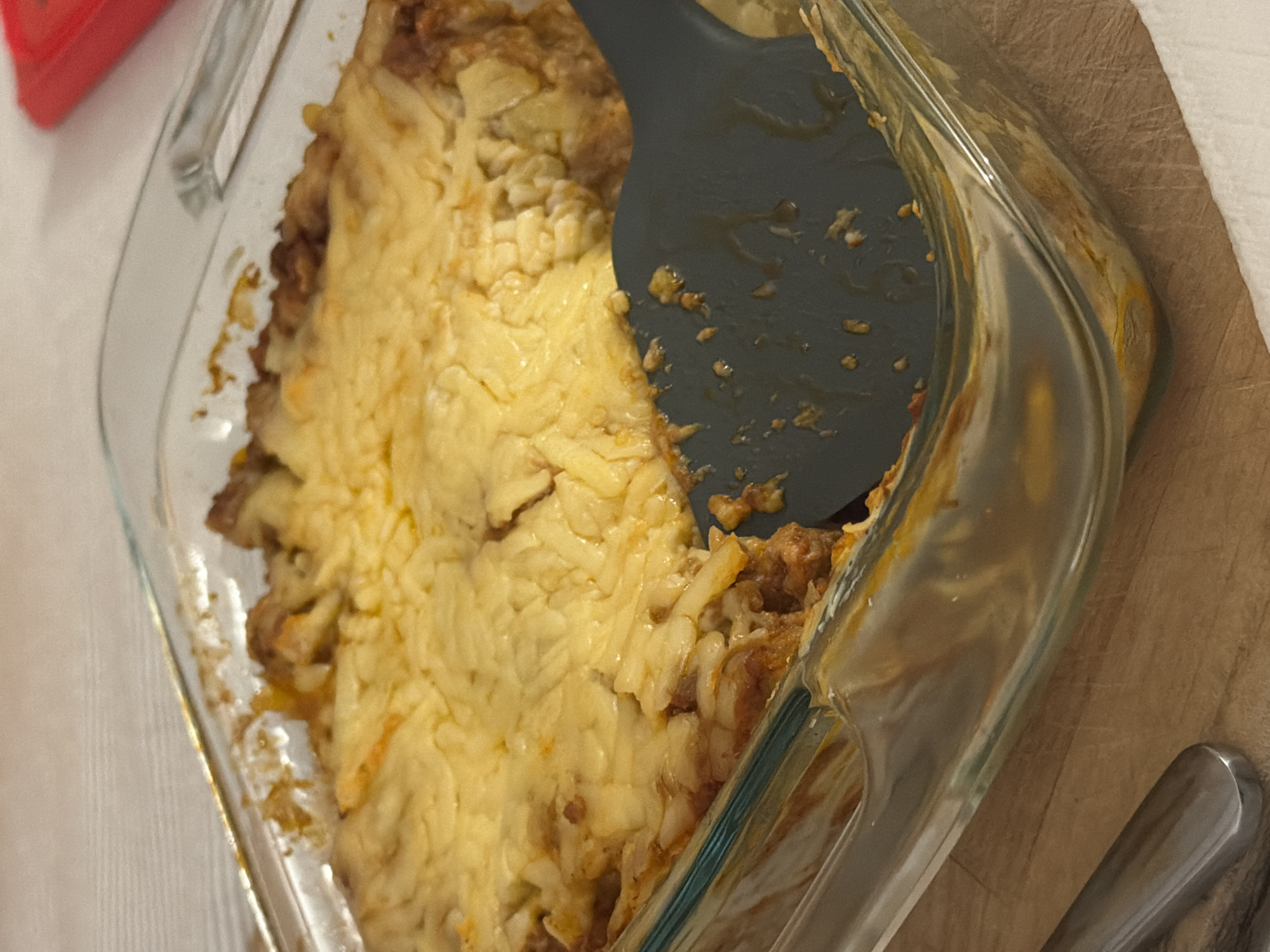 The image shows a glass baking dish filled with a food item that appears to be a casserole or lasagna. The top layer is covered with melted cheese, possibly cheddar or a similar variety. A dark gray spatula is partially inserted into the dish, with some of the casserole clinging to it. The dish is sitting on a wooden surface, likely a cutting board or countertop. A portion of a metallic object, possibly another utensil, is visible at the bottom right of the image. The location appears to be inside a residence, as indicated by the kitchen setting. There is no city or state information to provide a location.