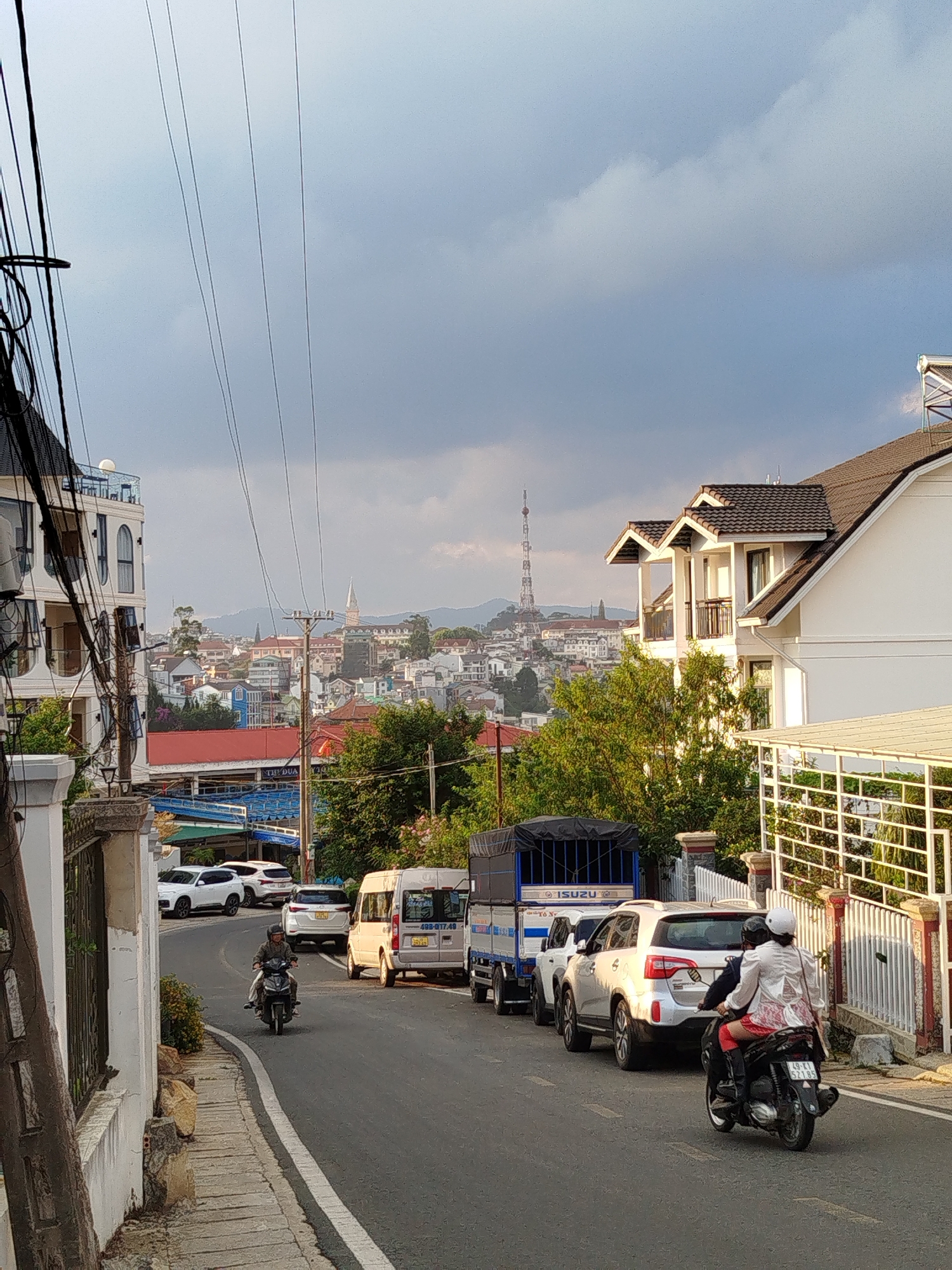 The image depicts a downward sloping street in Da Lat, Vietnam. The road is lined with parked cars and two motorcycles are traveling in opposite directions. The cityscape of Da Lat is visible in the background, including the Da Lat television tower. The sky is overcast with dark clouds. Buildings are present on both sides of the street, including multi-story structures and single-family homes. Power lines run across the upper portion of the frame. The street is paved and has a concrete sidewalk on the left side of the frame.