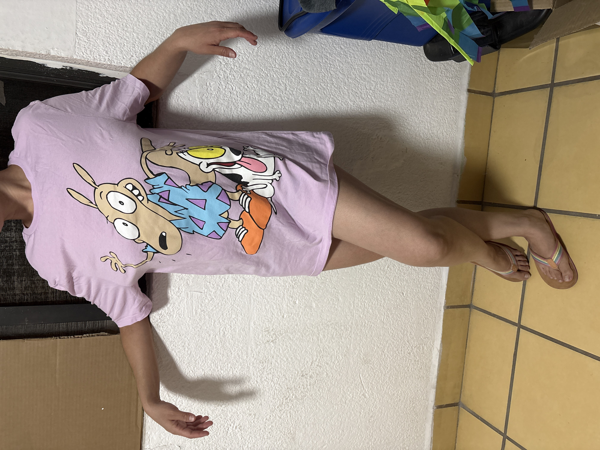 A person wearing a lilac t-shirt with a Rocko's Modern Life print and flip-flops stands on a yellow tile floor. The background consists of a textured white wall. A blue container and cardboard boxes are partially visible. Based on the image characteristics and the available information, there is no explicit evidence to confirm the location in the city of Cuernavaca, Mexico.