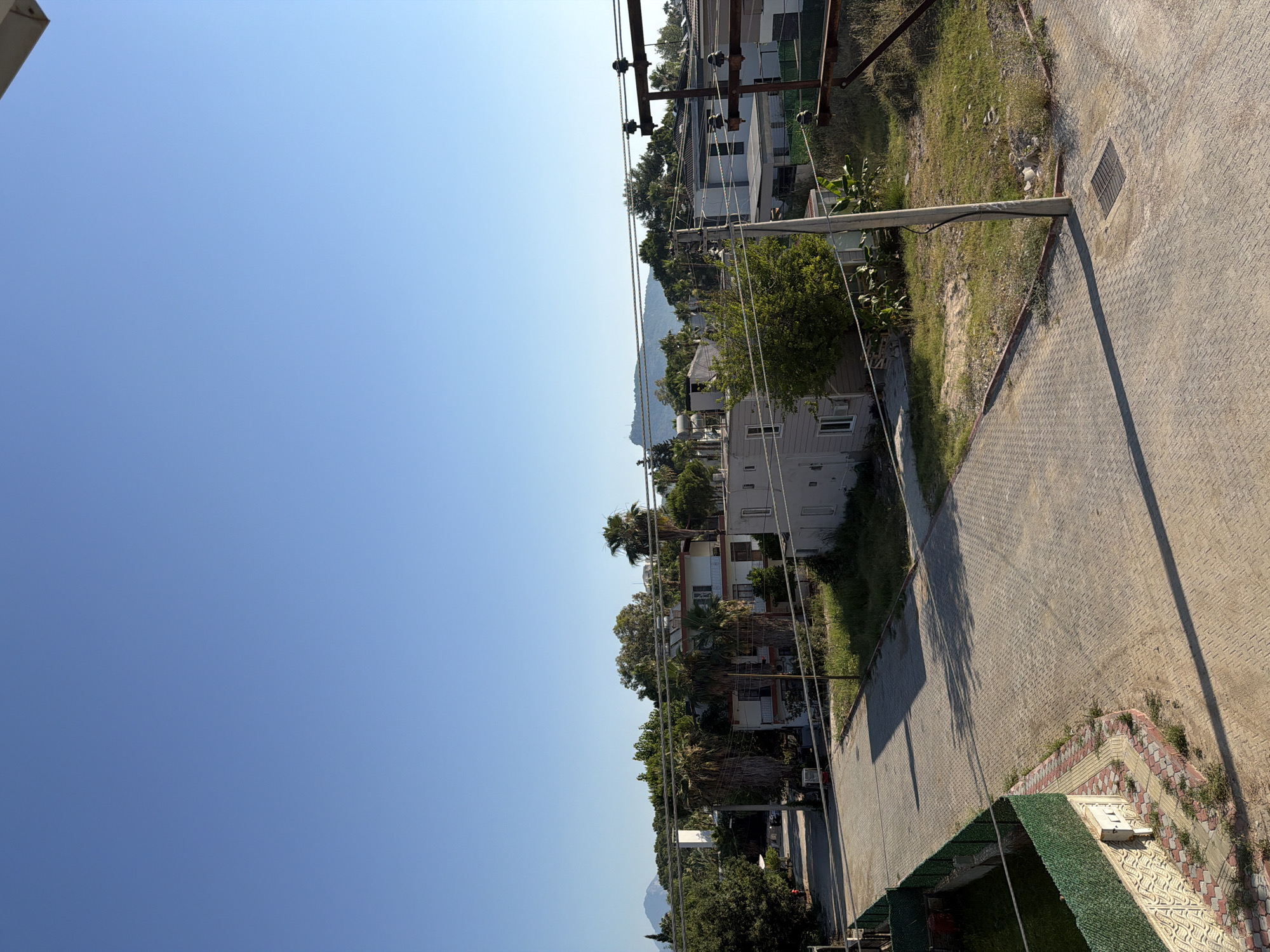 The media file depicts an outdoor scene bathed in bright sunlight, suggesting it was captured during the daytime. The sky is a clear, vibrant blue, indicating fair weather. The perspective is angled, looking down and to the right, across a paved area and then towards a row of buildings nestled amongst lush greenery.
The foreground is dominated by a textured, light-colored paved surface, possibly a sidewalk or road, with a dark grate visible on its surface. A shadow stretches diagonally across this paved area, cast by an unseen object or structure. To the left of the paved area, there is a strip of grass and some low-lying vegetation.
Beyond the paved area, a cluster of multi-story buildings can be seen. These appear to be residential or possibly small hotels, with white and cream facades and balconies. Palm trees and other leafy trees are interspersed between and around the buildings, creating a tropical or Mediterranean feel. In the distance, a gentle, tree-covered mountain range is visible under the bright sky.
Power lines and utility poles are prominent features, crisscrossing the scene and connecting the buildings. A concrete pole extends diagonally into the frame from the lower right.
The image does not contain any discernible people or text. The overall impression is one of a peaceful, sun-drenched day in a developed area with abundant natural vegetation. The orientation of the image is vertical, with the sky occupying the left portion and the built environment and landscape to the right.