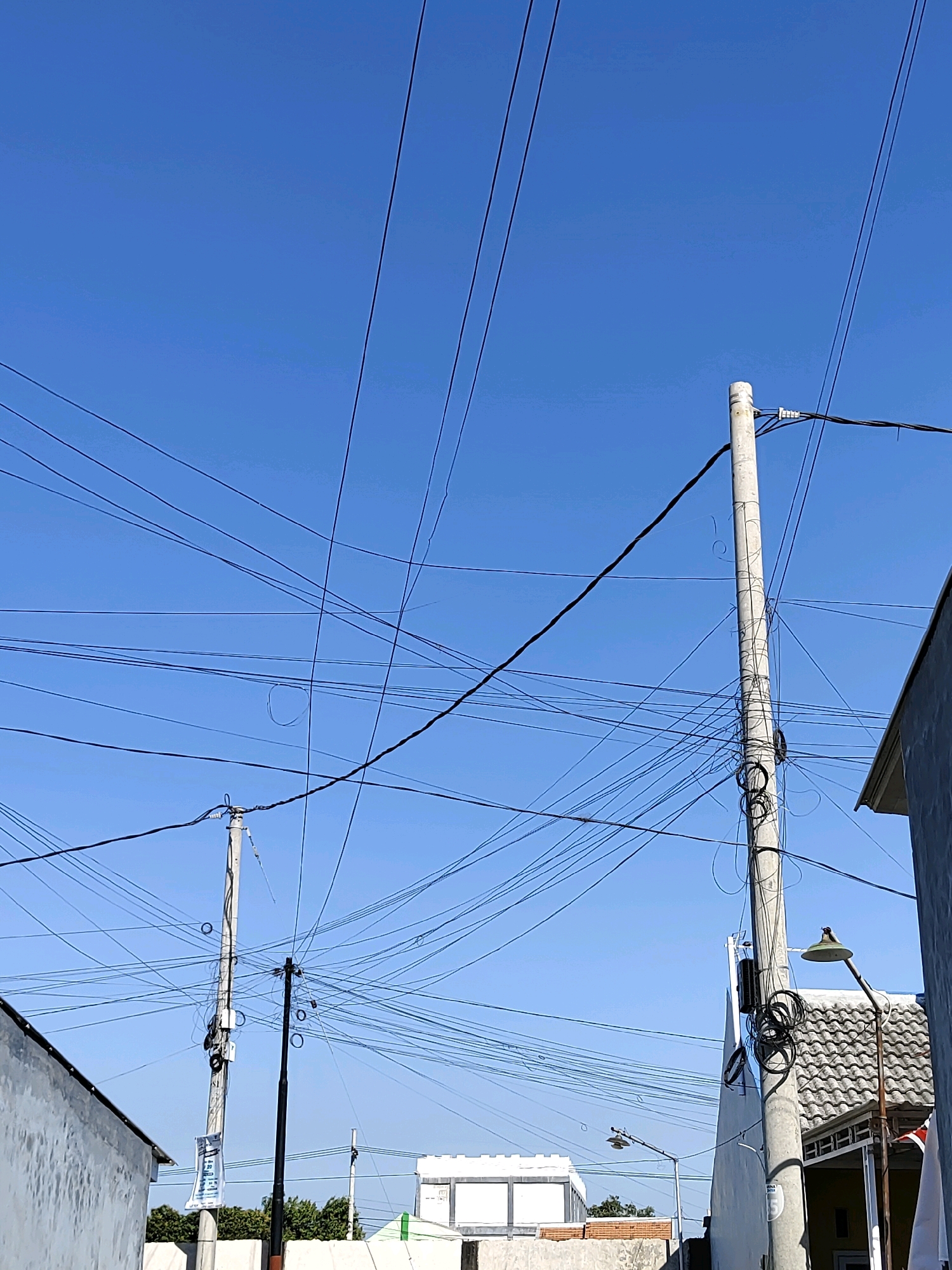 The image depicts a clear blue sky above a densely populated residential area, likely in Probolinggo, Indonesia, characterized by numerous overhead power and communication lines strung between poles. Two prominent concrete utility poles are visible, one taller than the other, with a profusion of black wires crisscrossing the frame. Some wires are taut, while others hang loosely, forming loops and tangles. Buildings with light-colored walls and grey tiled roofs are partially visible on the left and right edges of the frame, suggesting a close proximity of structures. Streetlights, one on a dark pole and another on a grey pole, are also present, as are some distant green trees and low-lying structures. The overall impression is one of an urban or semi-urban environment with a high density of electrical and telecommunication infrastructure.