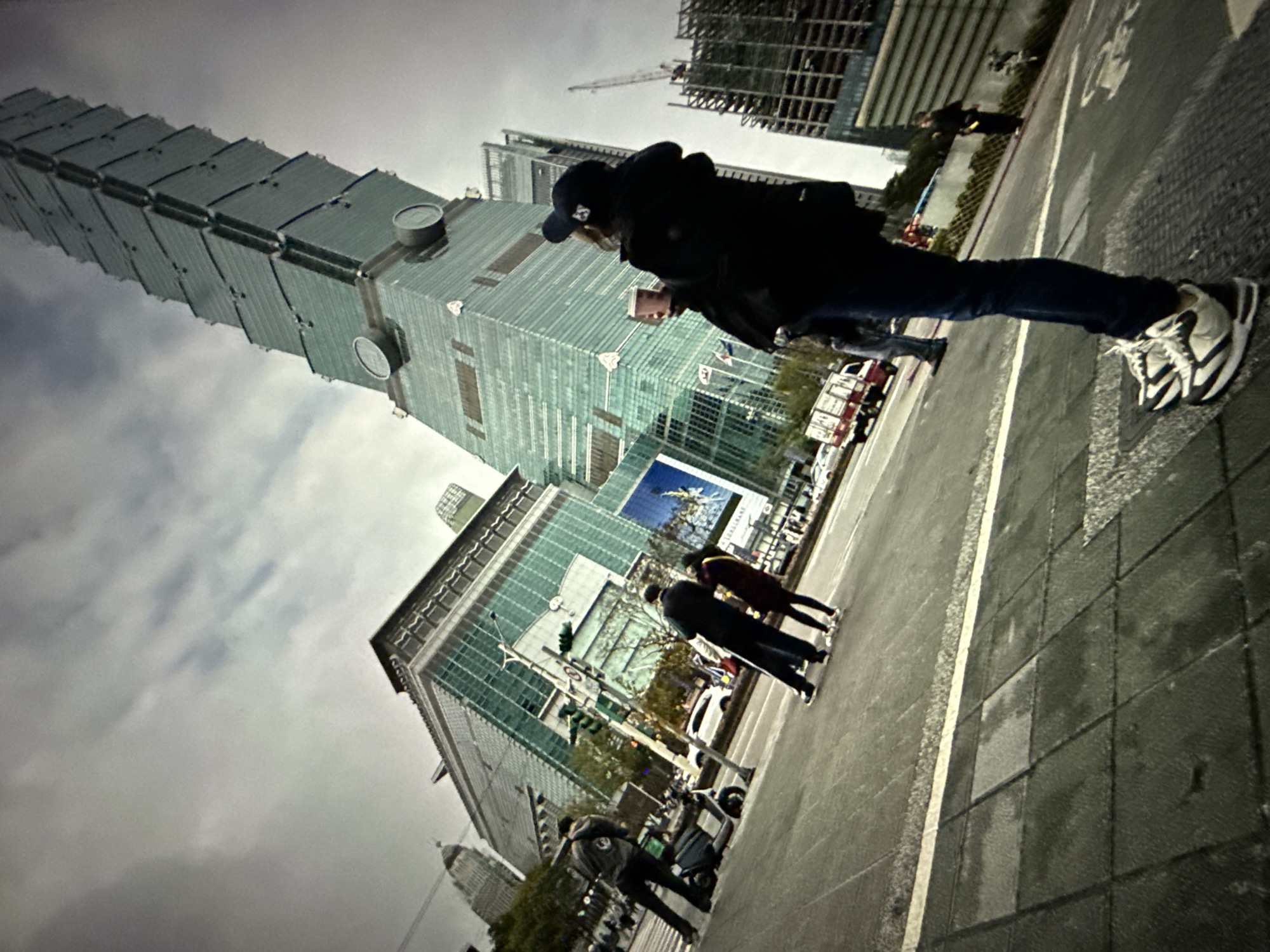 The image depicts a street scene in Taipei, Taiwan, with the iconic Taipei 101 skyscraper prominently featured. The sky is overcast, suggesting cloudy or possibly rainy weather. In the foreground, a person is standing, viewed from the waist down, wearing dark pants and white and black sneakers. They are holding a light-pink cup. Behind them, other individuals are walking and standing on a paved street with visible crosswalk markings. A scooter is parked to the left. In the mid-ground, several buildings are visible, including a large, modern glass-fronted structure and other lower buildings. Further back, the Taipei 101 tower rises into the sky, its distinctive segmented design visible. Construction cranes are also present, indicating ongoing development in the area. The overall impression is of a busy urban environment.