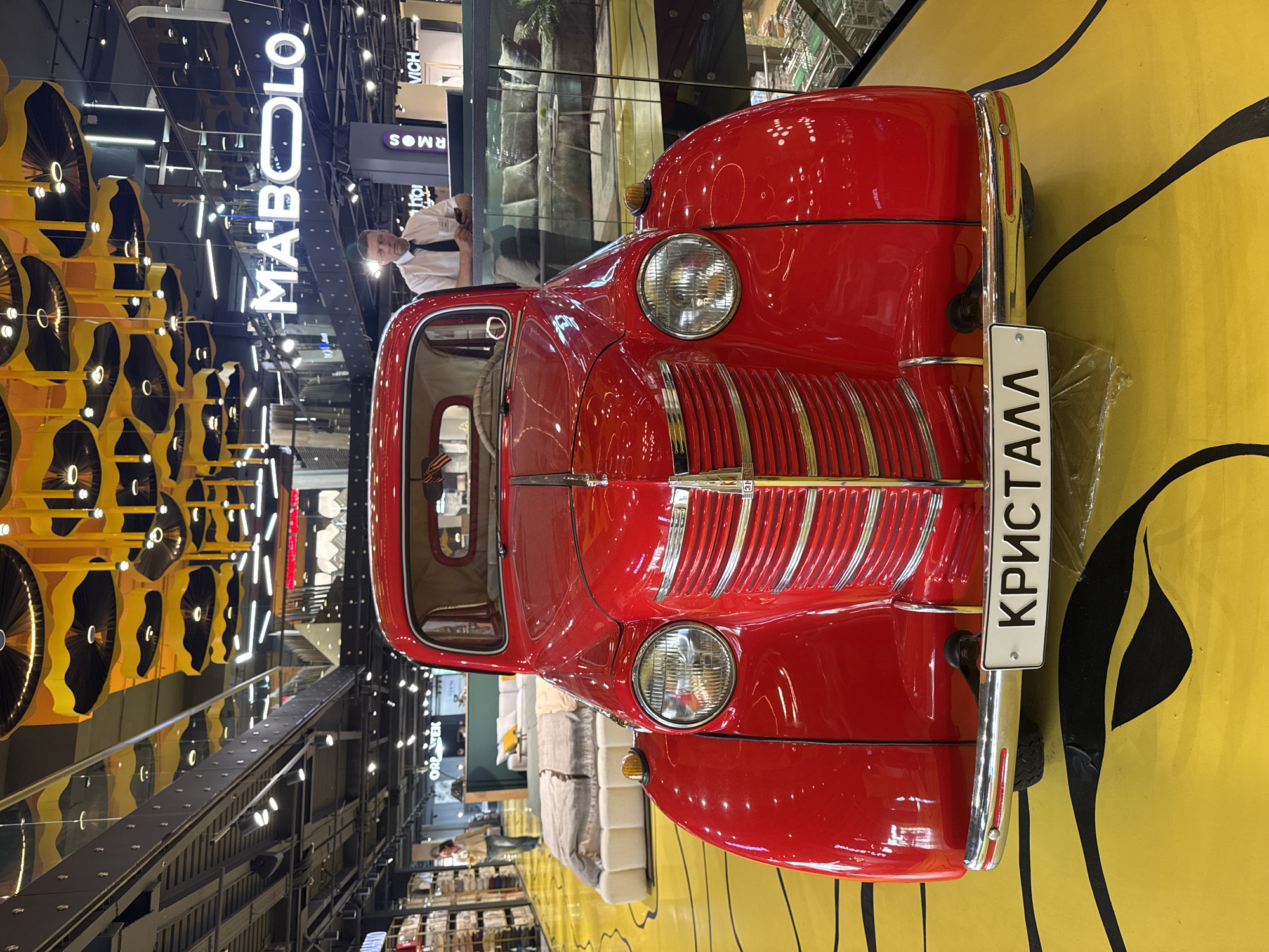 The image shows a red vintage car with a license plate reading "КРИСТАЛЛ" (KRISTALL) parked inside what appears to be a modern shopping mall. The mall setting is indicated by the presence of storefronts, illuminated signs (e.g., "MABOLO"), and other retail displays. In the background, a person in a white shirt and tie can be seen standing inside one of the storefronts. The floor is decorated with a striking yellow and black graphic pattern. The photo location is likely in Moscow, Russia, given the Cyrillic characters on the license plate. Additional interior decorative lighting and design elements suggest an upscale shopping environment.