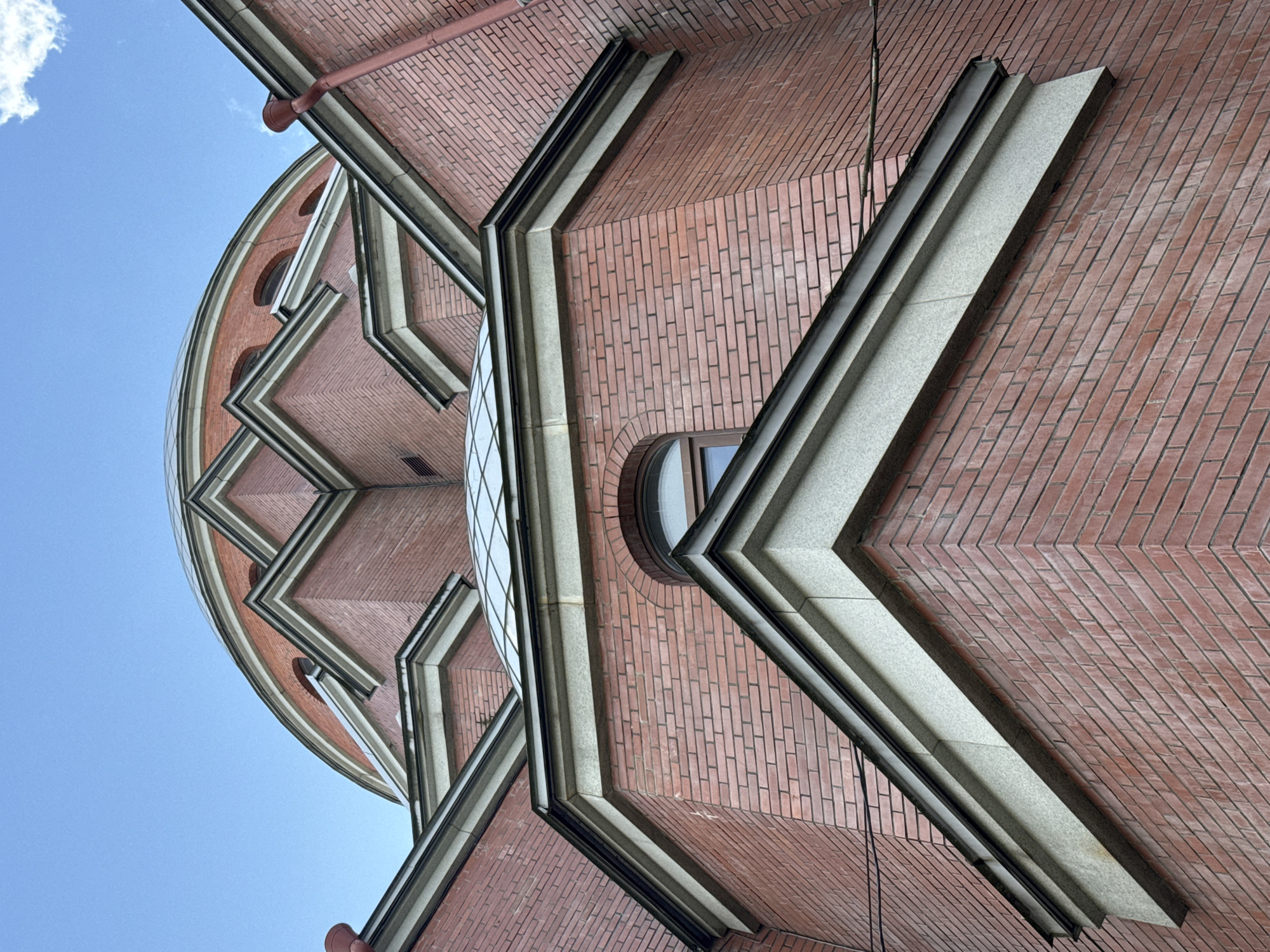 This is a low-angle exterior shot of a building with a red brick facade, featuring a domed roof and multiple angular roof sections. The building has several arched windows and decorative stone trim along the rooflines. A downspout is visible on the right side of the image. The sky above is blue with a few wispy clouds. The architectural style suggests it could be a religious building or a historical structure. The image is not specific to any particular location in Saint Petersburg without further information.