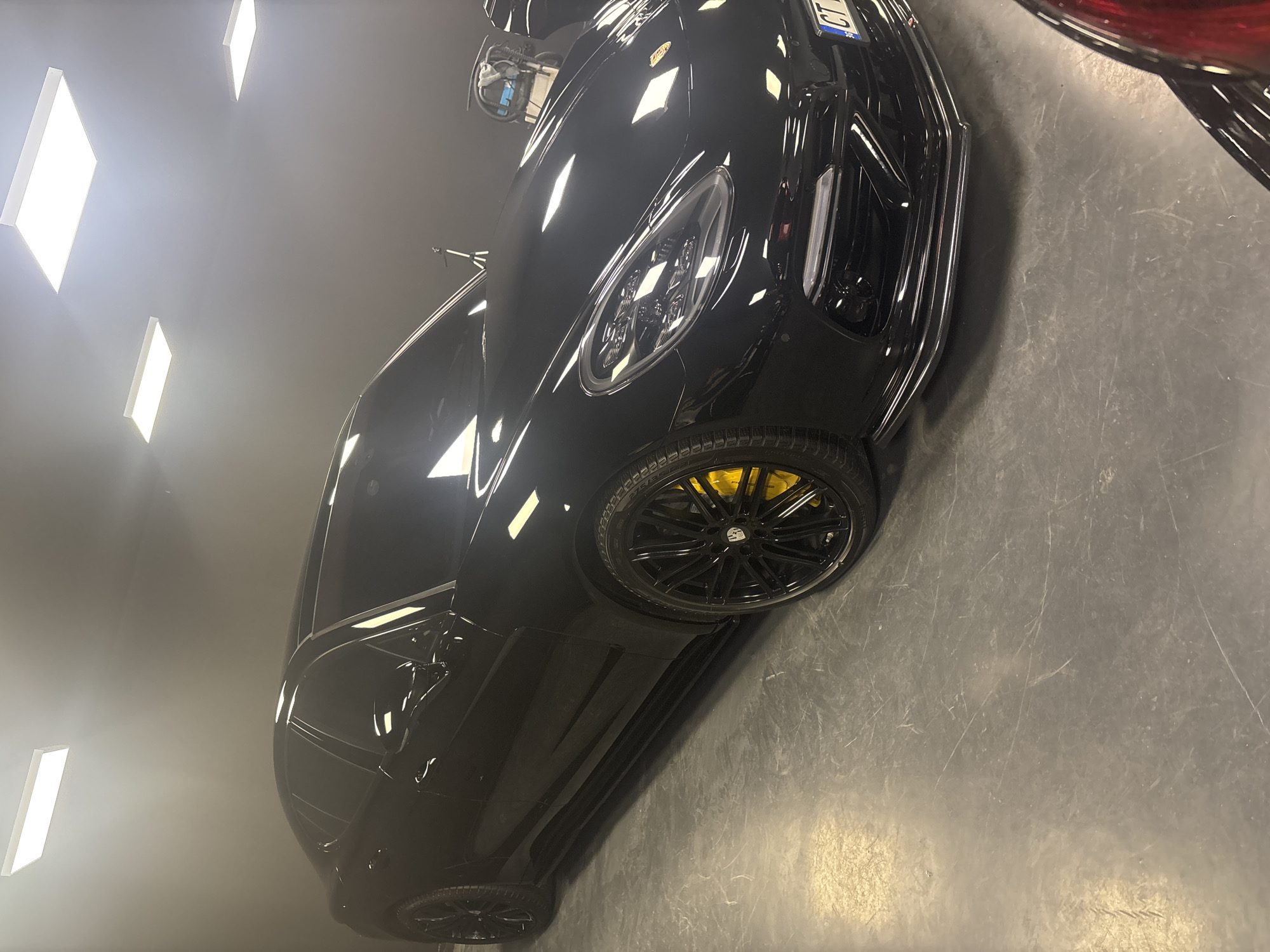 The image displays a highly polished black Porsche Panamera positioned indoors, likely within a detailing facility or specialized garage in Ruse, Bulgaria. The vehicle's sleek dark paint reflects the bright, rectangular LED panel lights on the dark grey ceiling. It features black multi-spoke wheels, which clearly reveal bright yellow brake calipers, and an aftermarket front lip spoiler. The front license plate, partially visible, reads "CT BG 30..." indicating its registration in Stara Zagora, Bulgaria. The floor is a grey concrete surface, showing multiple scuff marks and scratches. In the background, to the right of the car's front section, a silver tripod stands, and further back, a utility cart loaded with blue and white items, presumably detailing supplies, is visible. A sliver of another dark-colored vehicle is discernible in the bottom right corner of the frame. No individuals are present in the scene.