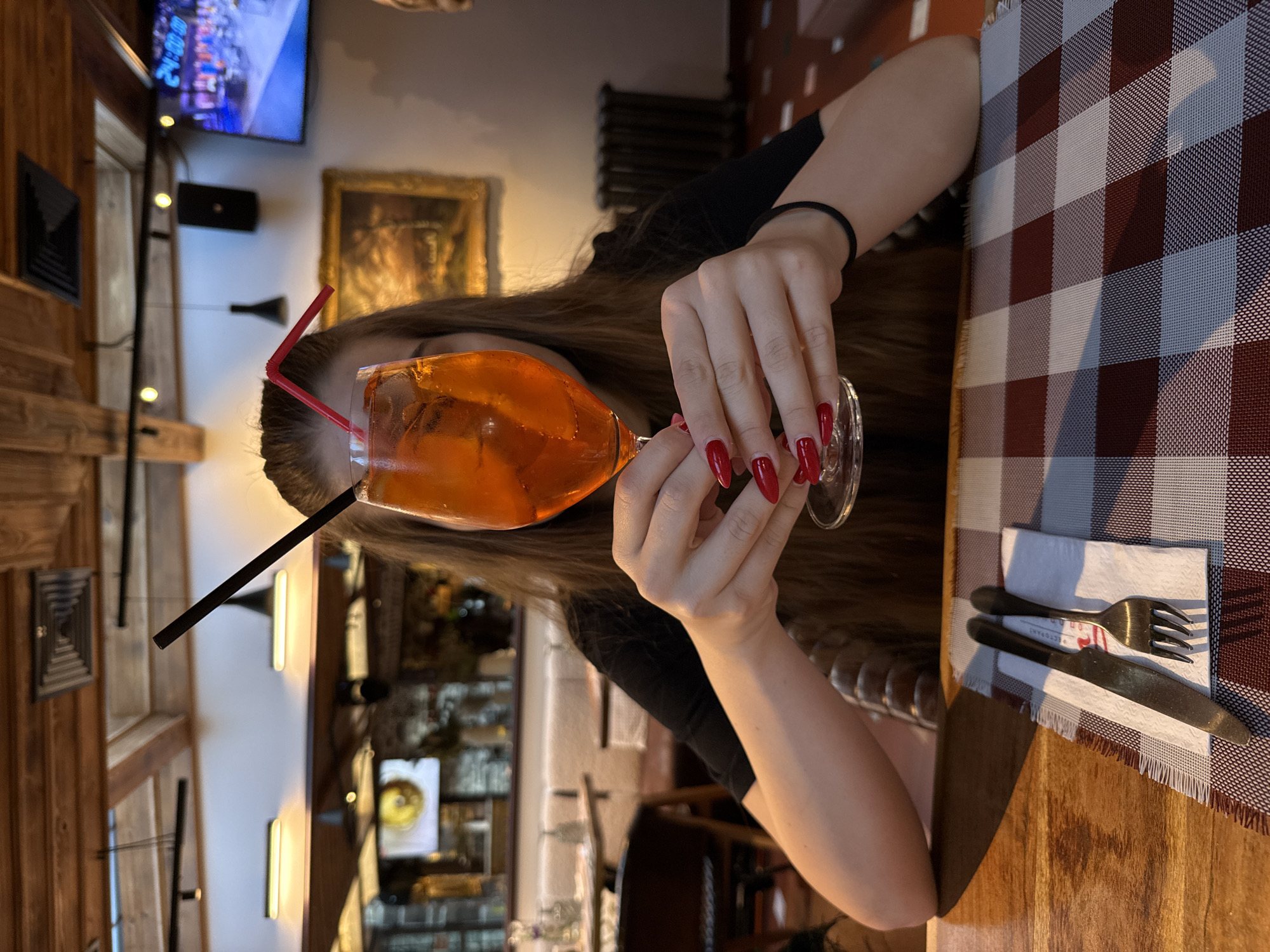 A woman with long, brown hair and bright red nail polish holds a glass of an orange-colored beverage with two straws, a black one and a red one, in a restaurant. She is wearing a black t-shirt and a black bracelet on her left wrist. The drink is in a large wine glass, filled with ice and liquid. In front of her on a wooden table, a red and white checkered placemat holds a white napkin with a silver fork and knife. The background shows a restaurant interior with exposed wooden beams on the ceiling, track lighting, and a painting on the wall. A television screen is visible in the upper right corner, displaying an image. The restaurant appears to be in the city of Shiroka Laka, Bulgaria.