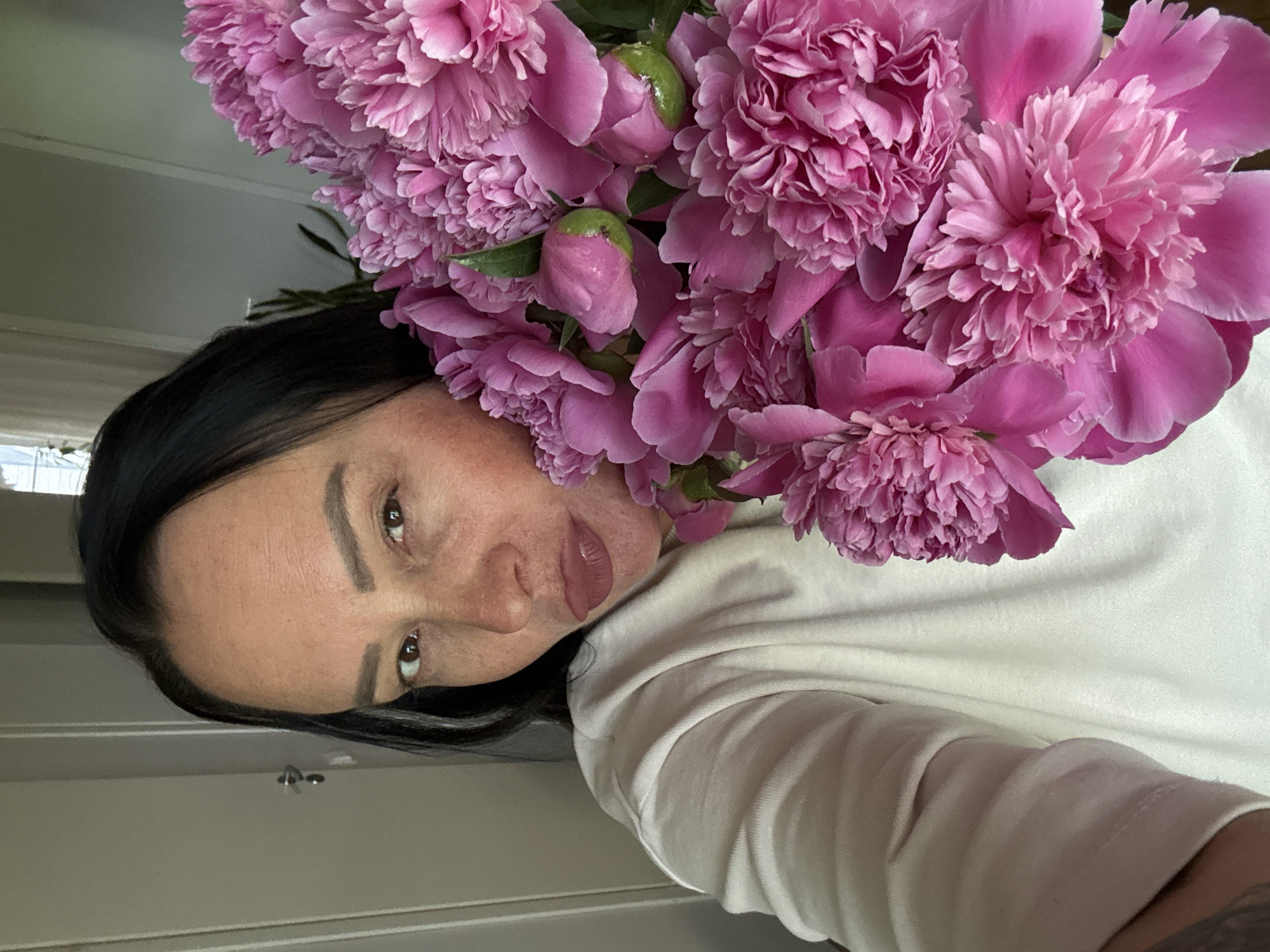A woman with dark hair and light skin poses with a large bouquet of pink peonies. She is wearing a white shirt. The background appears to be an interior space with a door. There is no information available in the image to identify the location in Chaikovsky, Russia.