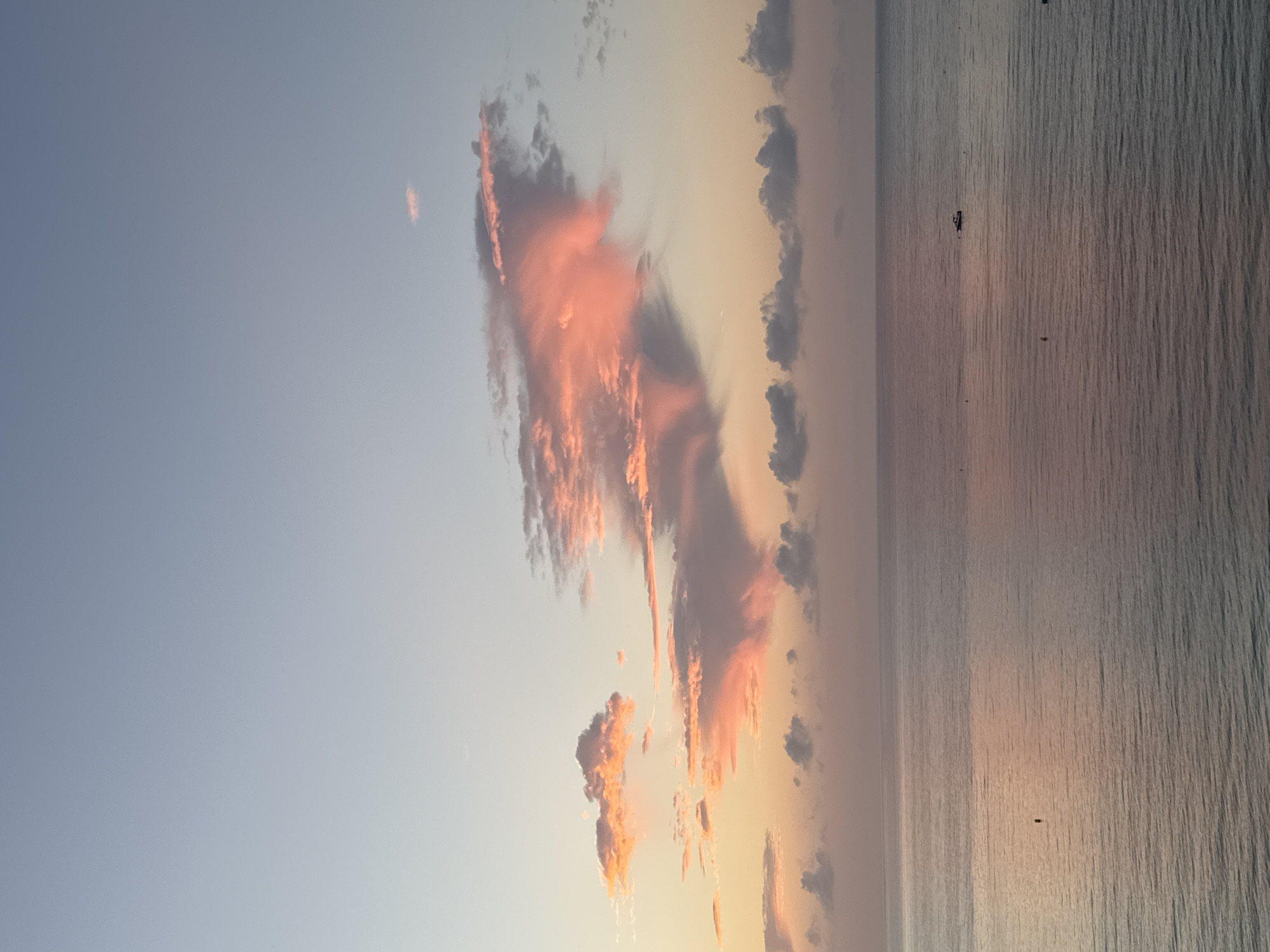 The image shows a serene view of the Mediterranean Sea at sunrise, likely from Calp, Spain, given the presence of the iconic Peñón de Ifach rock formation visible in the distance. The sky is a gradient of soft blues, pinks, and oranges, illuminated by the rising sun. Several clouds are scattered across the sky, catching the light and displaying vibrant shades of pink and coral. The sea is calm with gentle ripples reflecting the colors of the sky. A few small buoys and a boat are visible on the water, suggesting a peaceful morning. The overall atmosphere is tranquil and picturesque.