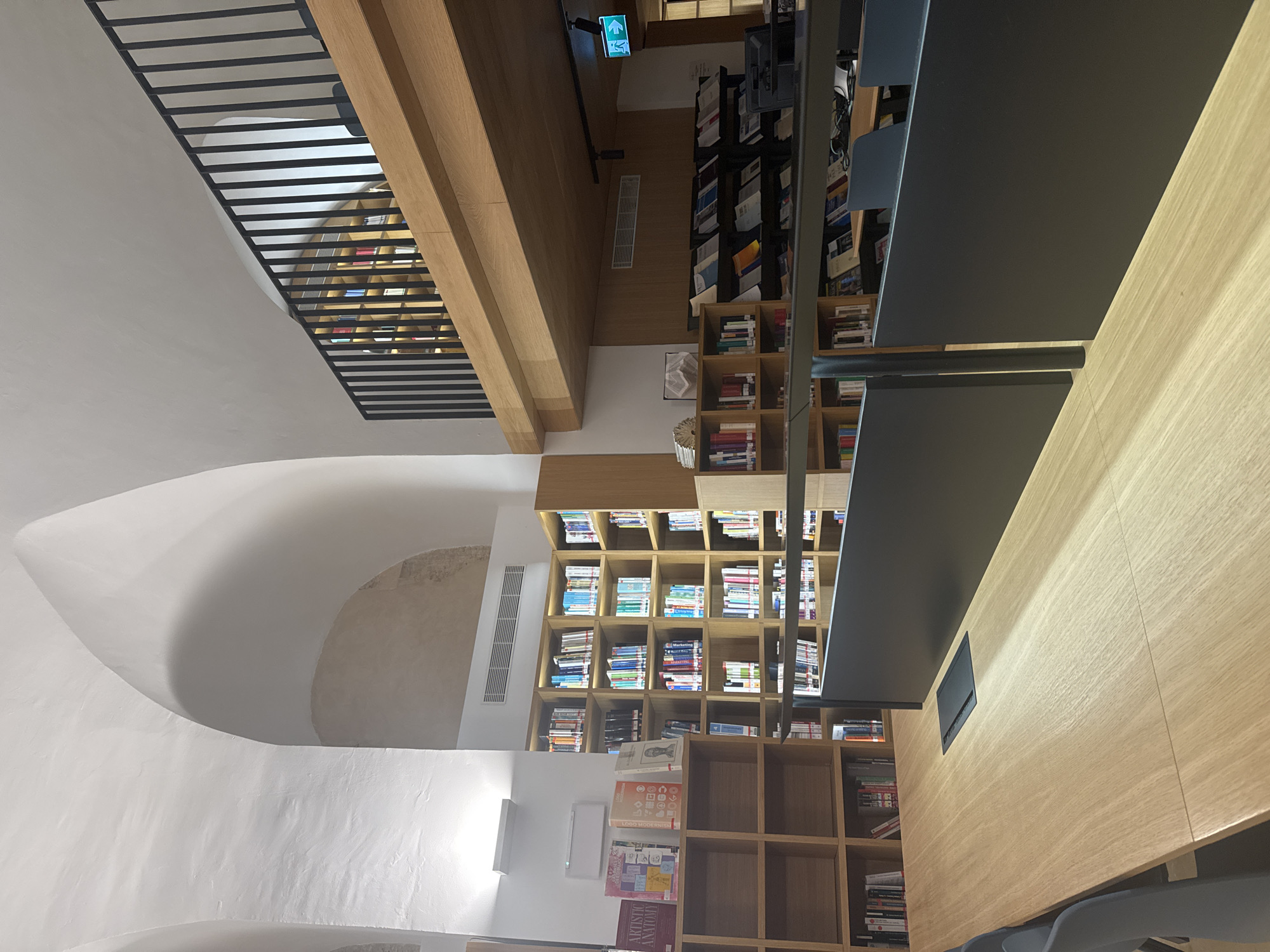 The image shows the interior of a library with high arched ceilings, a mezzanine level with a black railing, and rows of bookshelves filled with books. The library features wooden furniture, including tables and shelves, and has a modern aesthetic. An exit sign with a green running man symbol is visible on the wall. The scene appears to be in a public or academic library, possibly located in the city of València, Spain, given the context of the provided information. There are no people visible in the image.
