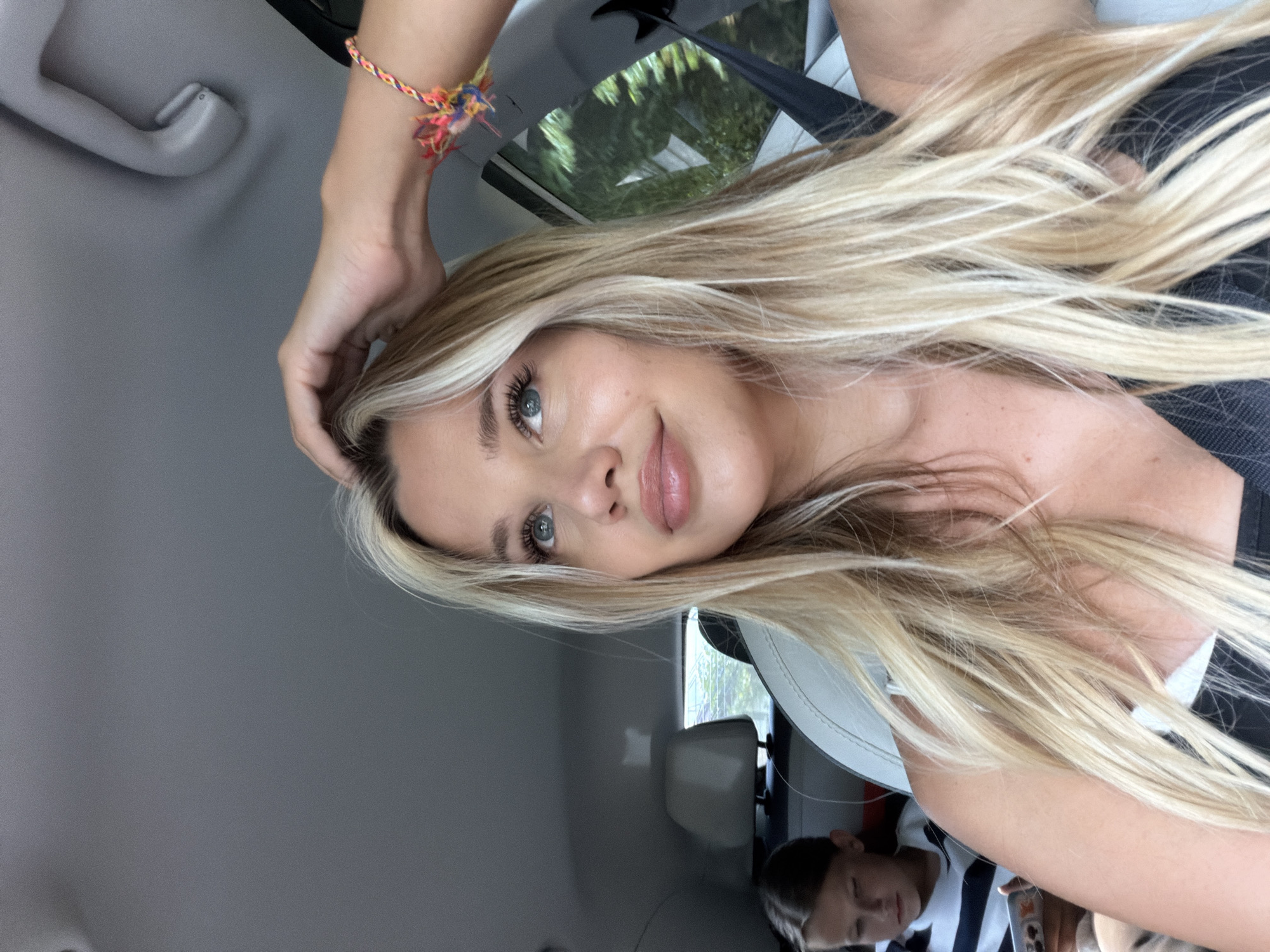 The image shows a close-up of a young woman with blonde hair and blue eyes, sitting in the passenger seat of a car. She is looking upwards and has her hand on her head. A young girl, likely a child, is visible in the back seat, looking down at a phone. The interior of the car is visible, including a light-colored headrest and the car's ceiling. The woman is wearing a black top and a colorful bracelet on her wrist. The scene appears to be during the daytime, with trees visible through the car window, suggesting an outdoor environment. There is no visible indication of the location being in the city of Wichit, Thailand.