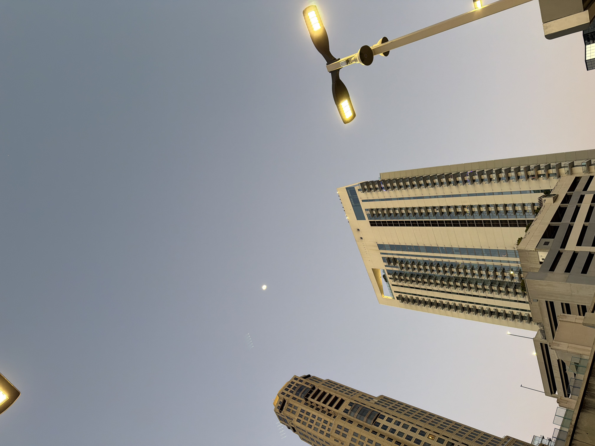 The image is a low-angle shot of the Atlanta skyline at dusk or dawn, featuring three buildings, a streetlight, and the sky. The building at the top right is a modern high-rise, primarily white with blue-tinted windows and numerous balconies. A second building at the bottom is older, constructed of yellow brick with a more ornate design. A third building on the right side of the frame is partially visible and appears to be a modern structure.
A streetlight with two illuminated lamps is situated in the upper part of the frame. The sky is a pale blue-gray, indicating twilight. A small, pale dot is visible in the sky which could be the moon or a star. The image appears to be taken from street level.