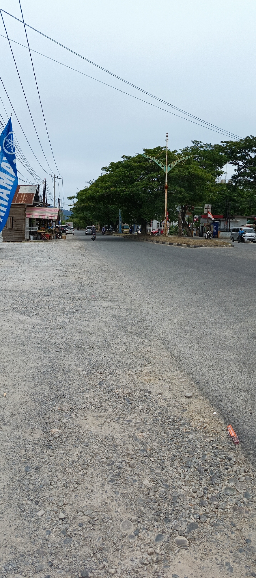The image depicts a street scene in Lampeuneurutblang, Indonesia, on an overcast day. In the foreground, a rough, gravel-covered area is visible, transitioning to an asphalt road. A small, rustic wooden stall with a corrugated metal roof is situated on the left side of the road, displaying various items, possibly for sale, with a blue Yamaha flag planted in front of it. Further down the road, a traffic intersection is present with several vehicles, including motorcycles, cars, and a truck. A street light with an ornate, possibly decorative, metal structure stands near the intersection. Lush green trees line the side of the road, and in the distance, a hilly landscape can be seen. The sky is predominantly gray, indicating cloudy weather. Several utility poles and power lines are strung across the scene. The overall impression is of a moderately busy local road in a tropical or subtropical region.