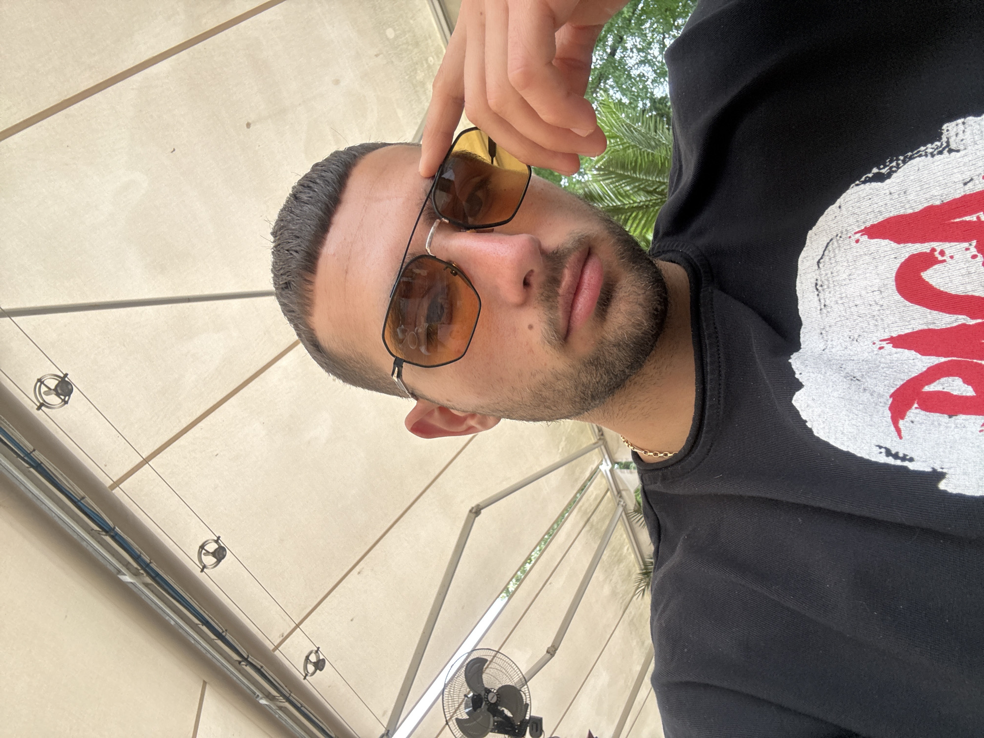 A medium shot photograph depicts a man wearing tinted glasses and a black sweatshirt. The scene is illuminated by overhead lighting. The location is suggested to be outdoors under a structure with electrical wiring and a fan, potentially in Ruse, Bulgaria. The sweatshirt has a partial white and red graphic. The man is holding his glasses with his hand.