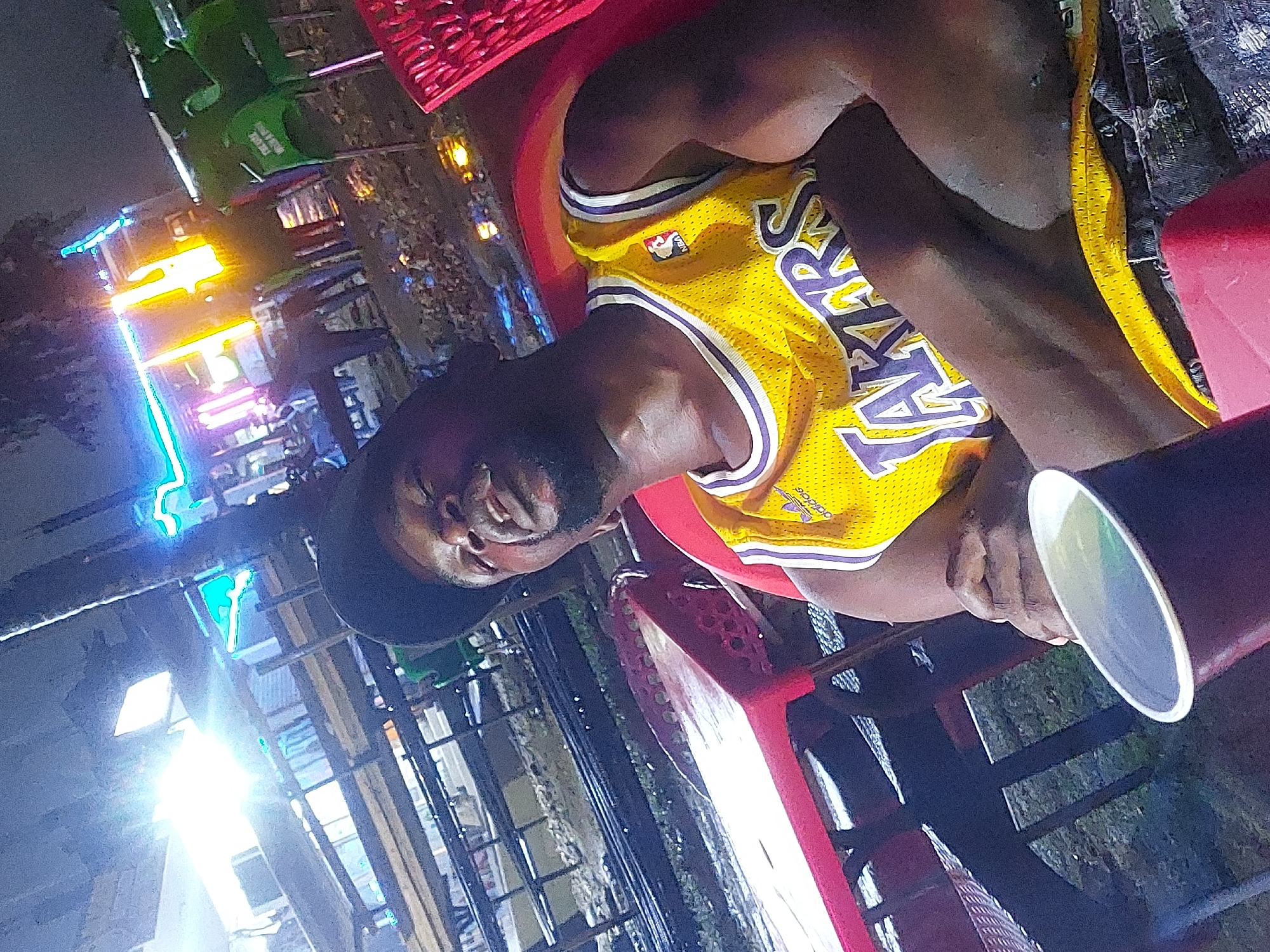 An outdoor, nighttime image, taken from a tilted angle, features a Black man smiling or laughing, looking towards the camera. He is wearing a yellow Los Angeles Lakers basketball jersey with purple lettering ("LAKERS") and the NBA logo visible on the chest. The "adidas" logo is also visible on the shoulder strap. He wears a dark hat and is seated on a red chair. In his right hand, he holds a dark-colored cup with a white rim.
The background suggests a vibrant, illuminated outdoor setting, possibly in Dar es-Salaam, Tanzania. Various bright, artificial lights dominate the scene, including prominent blue, green, and yellow/orange neon lights or similar fixtures. Some darker structures and hints of greenery or foliage are also visible behind him. The atmosphere appears lively and casual, suitable for an evening out at a market, outdoor eatery, or entertainment venue. The overall impression is one of enjoyment in a bustling nocturnal environment.