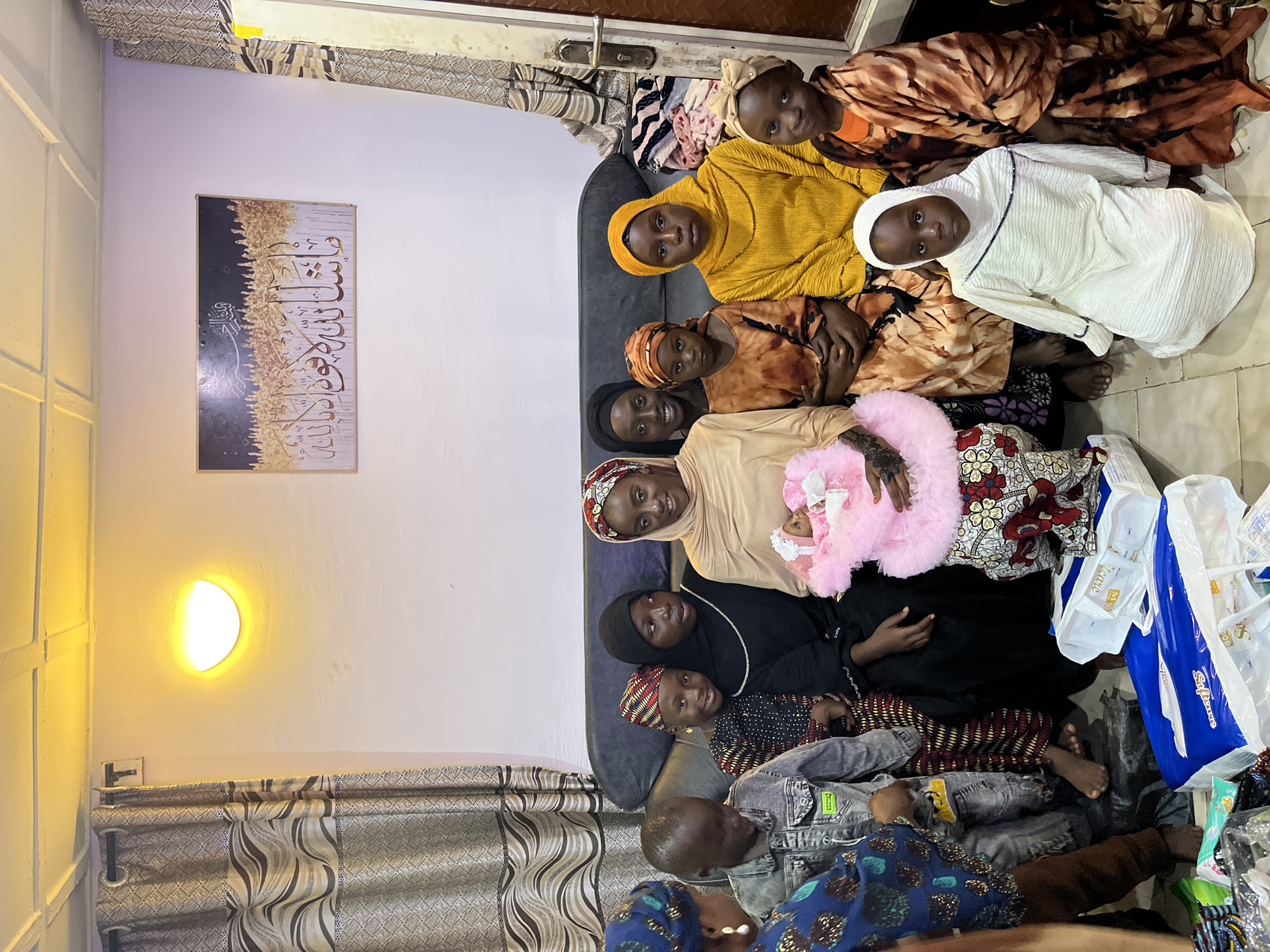 A photograph captures a gathering of ten individuals, primarily children and women, inside a residential setting. The scene appears to be an indoor living room, indicated by the presence of a couch, curtains, and wall decor.
The group is positioned on and around a dark-colored couch, with some standing behind and others sitting. The individuals are dressed in a variety of attire, predominantly traditional Islamic clothing, including hijabs and patterned fabrics. One child is holding a doll dressed in pink.
Prominently displayed on the wall behind the group is a framed piece of Islamic calligraphy art, adding to the cultural context of the scene. To the left of the frame, a patterned curtain is visible, and a wall-mounted light fixture emits a warm glow, illuminating the space. The floor is tiled.
The image does not contain any elements that specifically identify the location as Jos, Nigeria.
