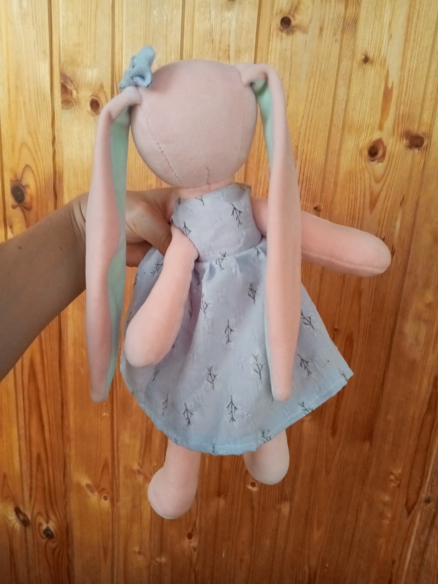 A handmade plush rabbit toy is held by a person's arm against a wooden wall. The rabbit is pink and has long, floppy ears, one with a light blue bow. It wears a light blue dress with a repeating pattern of small, dark trees. The wooden wall consists of vertical planks with visible knots and grain. The scene appears to be indoors, possibly in a residential setting in Chistopol, Russia.