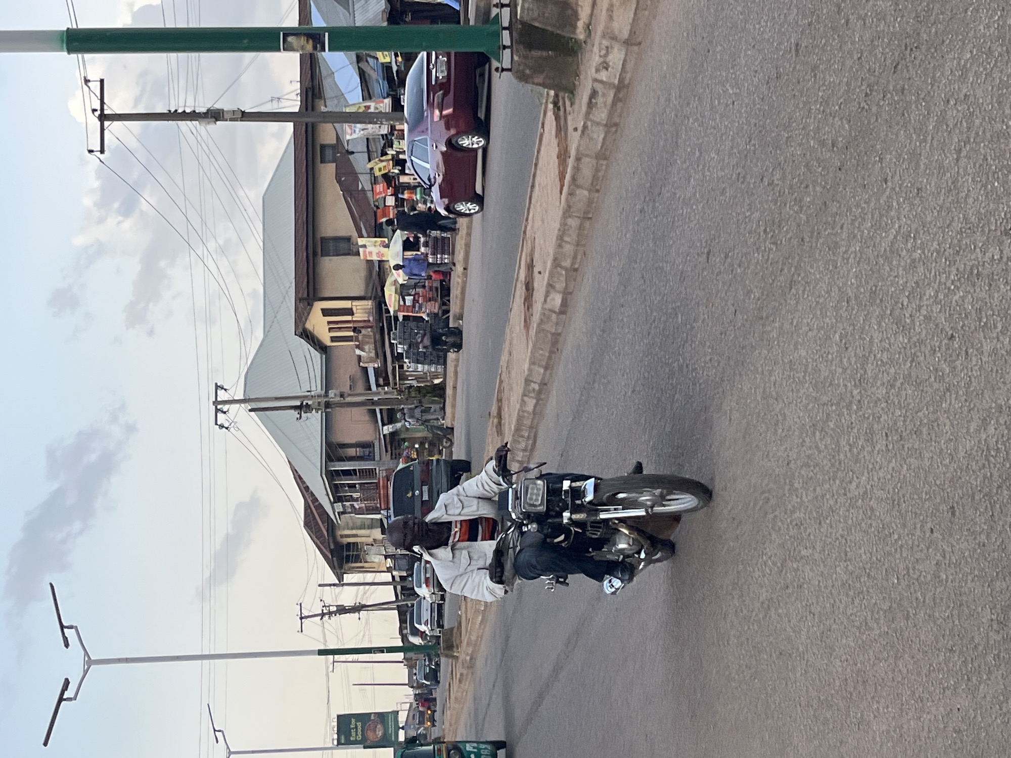 An outdoor, eye-level, medium shot captures a man riding a motorcycle down a paved road in an urban environment. The man, wearing a white jacket over a striped shirt and blue jeans, is the central focus of the image. He is looking forward, with his hands on the handlebars of his motorcycle. The motorcycle has a silver frame and a black seat, with a visible headlight and front wheel. The road is textured with asphalt and bordered by a concrete curb.
In the background, several buildings with corrugated metal roofs are visible, suggesting a commercial or residential area. A dark red sedan is parked on the right side of the road. Power lines and utility poles stretch across the sky, indicating an established infrastructure. The sky is partly cloudy, with a pale hue, suggesting it might be late afternoon or early morning. Other vehicles, including a green tricycle and several cars, are present on the road, indicating ongoing traffic. The scene appears to be in a bustling area with signs of commerce and daily life.