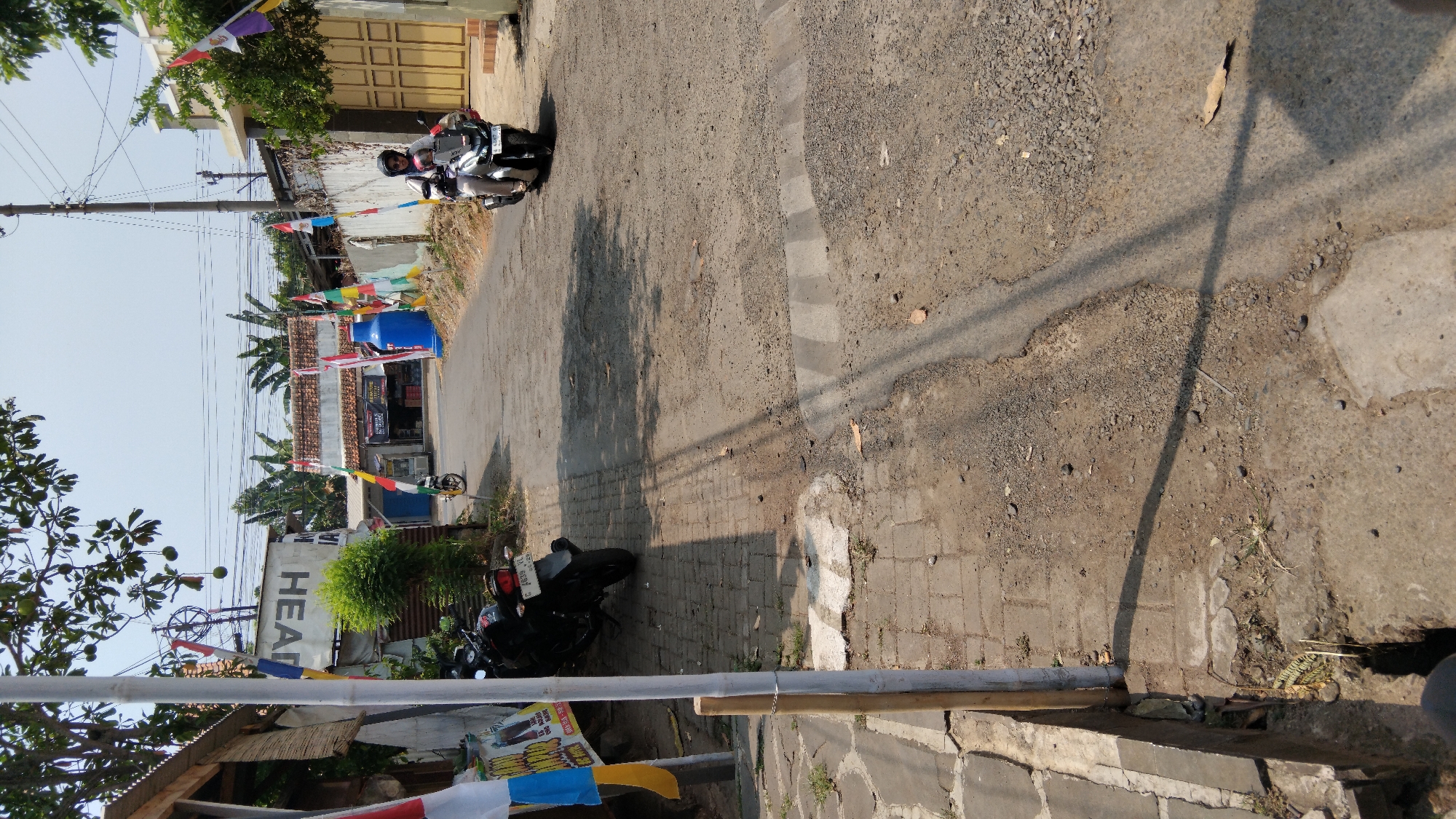 In the city of Pasekaran, Indonesia, a rural road is depicted under clear skies. The road is paved with asphalt and has a textured surface, indicating some wear. Several parked motorcycles are visible, including one in the foreground on the left side of the road, and another further down on the right, ridden by a person wearing a helmet and dark clothing. The road is lined with low-rise buildings and fences, some adorned with Indonesian flags. A large blue cylindrical object stands near one of the buildings. The environment appears to be a mix of residential and commercial areas, with some greenery present. Shadows are cast across the road, suggesting it is daytime.