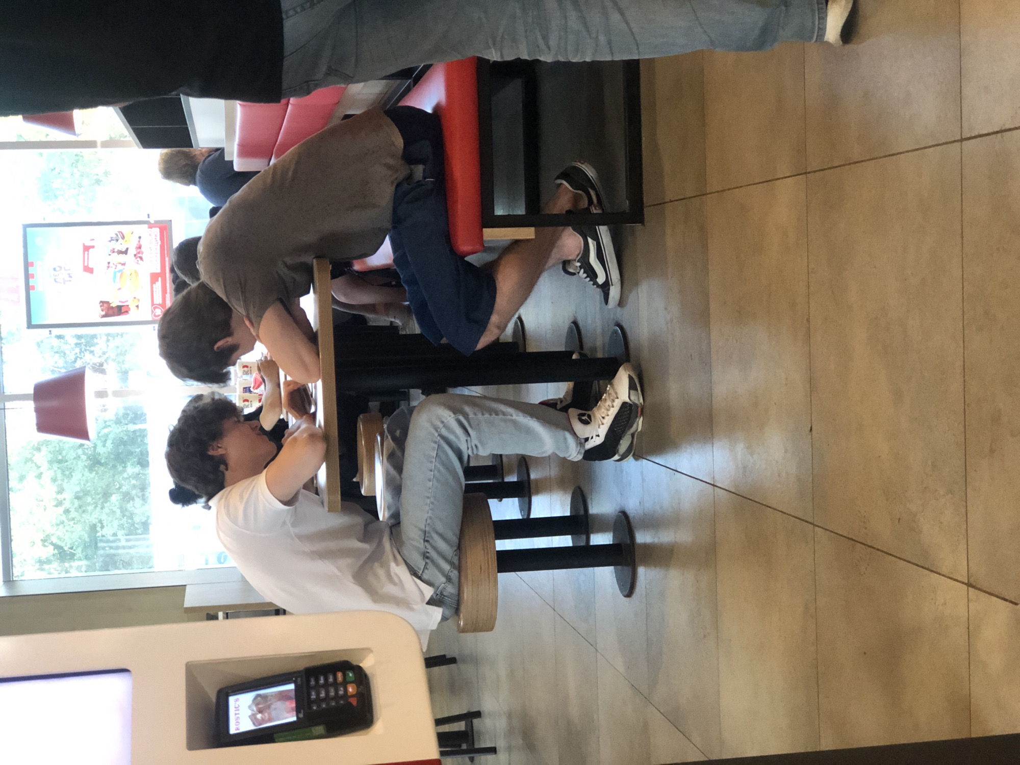 The image depicts an indoor scene within a fast-food restaurant located in Sestroretsk, Russia, during daytime. In the immediate foreground on the left, a light-colored kiosk features a black payment terminal with a numeric keypad and a color screen displaying the brand name "ROSTIC'S" and an image of food, consistent with a quick-service restaurant environment.
Two young males are seated at a wooden table in the center. The male on the left, wearing a white short-sleeved t-shirt, light blue jeans, and white, black, and red sneakers, is seated on a round wooden stool. He leans forward with his forearms resting on the table, directing his gaze downwards. Opposite him, another young male, wearing an olive green t-shirt, dark blue shorts, and black Vans Old Skool sneakers with white stripes, is seated on a red upholstered bench. He also leans forward with forearms on the table, engaging with the space between himself and the other individual. Both appear focused on something positioned on the table surface.
In the background, other patrons are visible, along with additional red upholstered bench seating. Through a large window behind the seated individuals, outdoor greenery and a bright sky are visible. A large poster featuring cartoon characters and text, including a red bucket logo typical of KFC and parts of Russian text, is visible through the window. Two red industrial-style pendant lamps hang from the ceiling. The floor is composed of light-toned square tiles with visible grout lines. A person's lower legs, clad in light blue jeans and white shoes, are partially visible in the right foreground, indicating someone standing nearby.