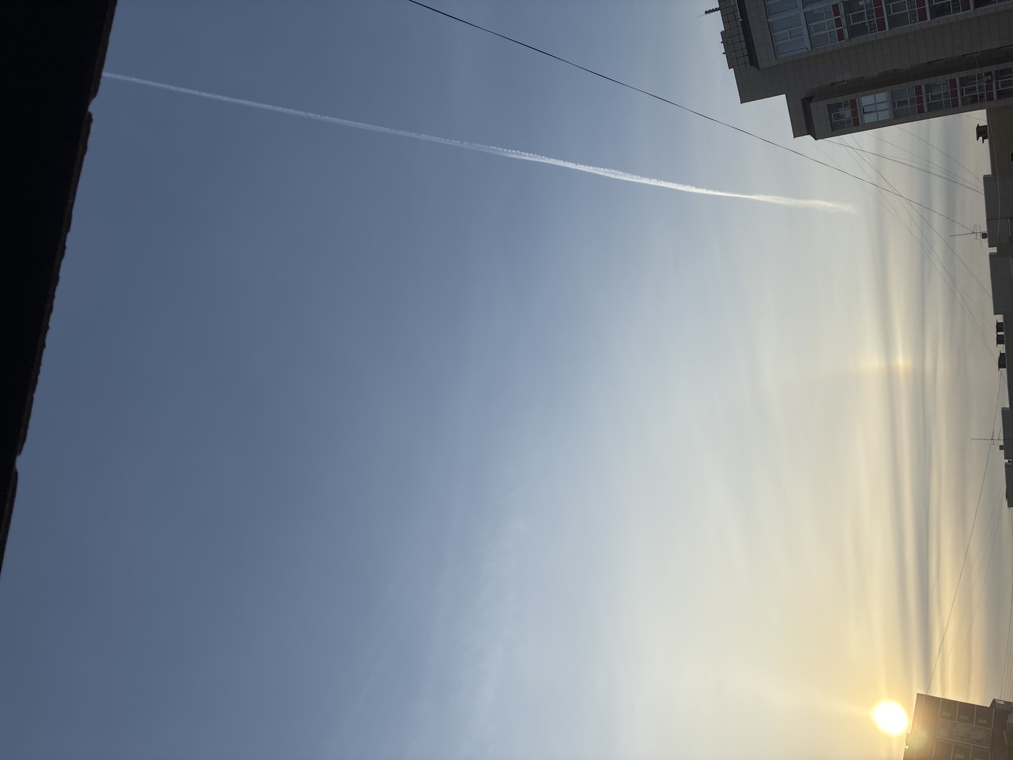 The image captures a bright, hazy sky, heavily tilted. The sky transitions from a deep blue on the left to a brilliant, glowing yellowish-white on the right, where the sun is prominent. A long, thin white contrail, presumably from an airplane, diagonally traverses the sky from the upper left to the mid-right.
On the far left, a dark, out-of-focus structure, possibly a wall or roofline of a building, creates a stark border. On the far right, the upper sections of a modern-looking, light-colored building are visible. It features several windows, some with distinct red frames, and what appear to be balconies or architectural details.
The sun, a very bright and almost perfectly circular orb, is situated in the bottom right corner, emitting an intense glare that stretches vertically across the right side of the sky, creating noticeable light streaks or atmospheric effects. Several thin, dark lines, likely electrical or communication wires, are strung across the upper-right portion of the frame, originating from near the building.
The scene depicts a clear day with strong sunlight, possibly morning or late afternoon, given the intense glow and atmospheric haze around the sun. There are no people visible, nor is there any discernible text.