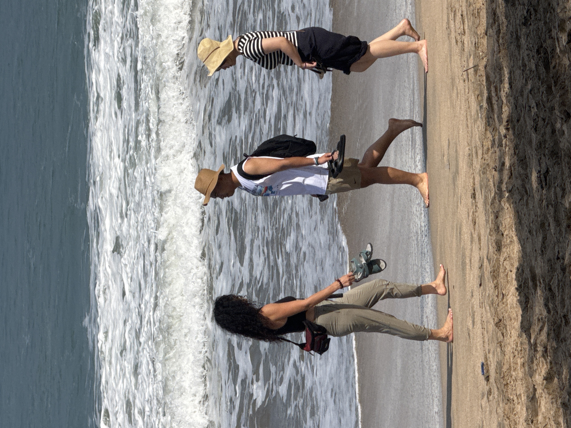 Three people are walking on the beach near the water's edge in Kuta, Indonesia. The person on the left, a woman with long dark curly hair, is wearing a black tank top, olive green pants, and a red and black fanny pack. She is holding a pair of dark sandals in her right hand and a pair of lighter sandals in her left hand. The person in the middle is a man wearing a white sleeveless t-shirt with a graphic print, khaki shorts, a tan fedora hat, and a black backpack. He is holding a pair of black flip-flops in his right hand. The person on the right is a woman wearing a black and white striped t-shirt, a dark skirt, and a light-colored sun hat. She is walking barefoot, holding a pair of dark sandals in her right hand. The ocean waves are breaking on the sandy beach, and the water appears to be a murky blue-green. The lighting suggests it is daytime.