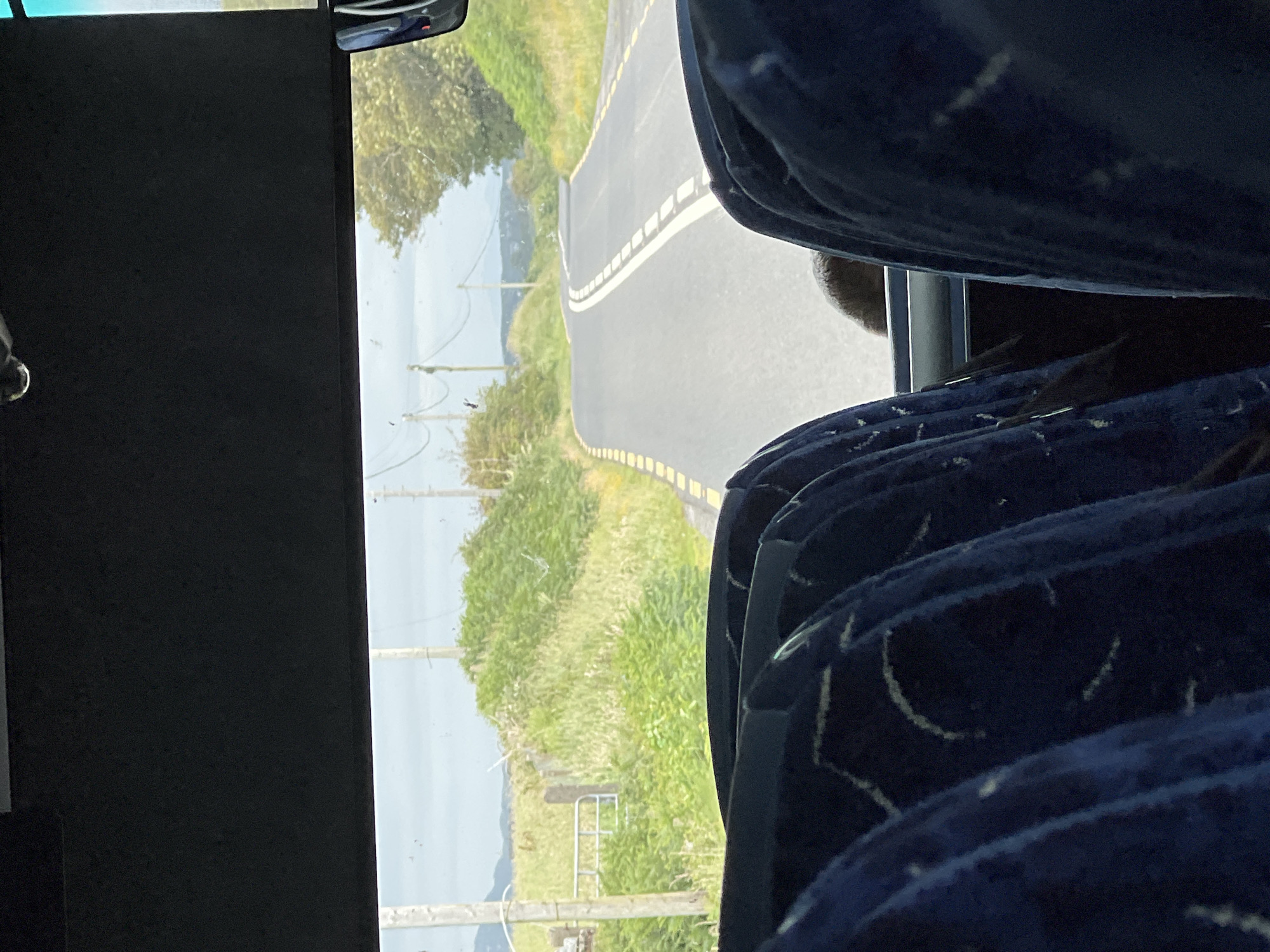 The image shows a view from the inside of a bus looking out at a winding road in Castlebane, Ireland. The foreground is filled with the backs of blue bus seats, some with white patterned stitching. In the middle ground, a paved road curves to the right, bordered by yellow and white dashed lines and surrounded by green grass and bushes. In the background, utility poles and wires are visible against a slightly overcast sky. Further in the distance, rolling hills and trees can be seen. A metal gate is also visible on the left side of the road. A person's head and shoulders, with brown hair, are partially visible between the bus seats.