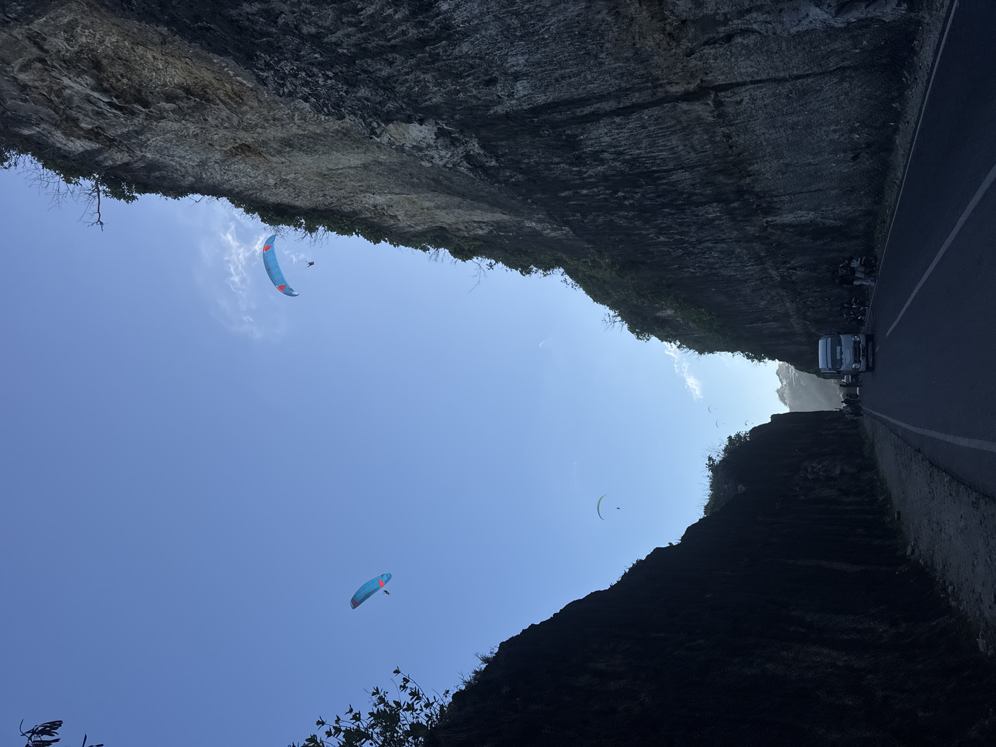 The image captures a scenic view along a road at the base of a towering cliff face, likely in Kuta Selatan, Indonesia, a region known for its dramatic coastal geology. The scene is dominated by the imposing, dark, textured cliff on the left, with the lighter, more weathered cliff face on the right. A paved road with a white line marking runs through the center of the image, with a dirt shoulder on the left.
Several paragliders are visible in the bright blue sky, soaring above the cliffs. One paraglider, with a blue and red canopy, is positioned high and to the right. Another, with a similar blue and red canopy, is closer to the center and lower. A third paraglider with a pale green canopy is visible further in the distance.
On the road, a white truck is present, and several motorcycles are parked or stopped along the side. A few individuals are gathered near the motorcycles and the truck. The foliage on top of the cliffs suggests a tropical environment. The overall impression is one of an adventurous and striking natural landscape integrated with human activity.