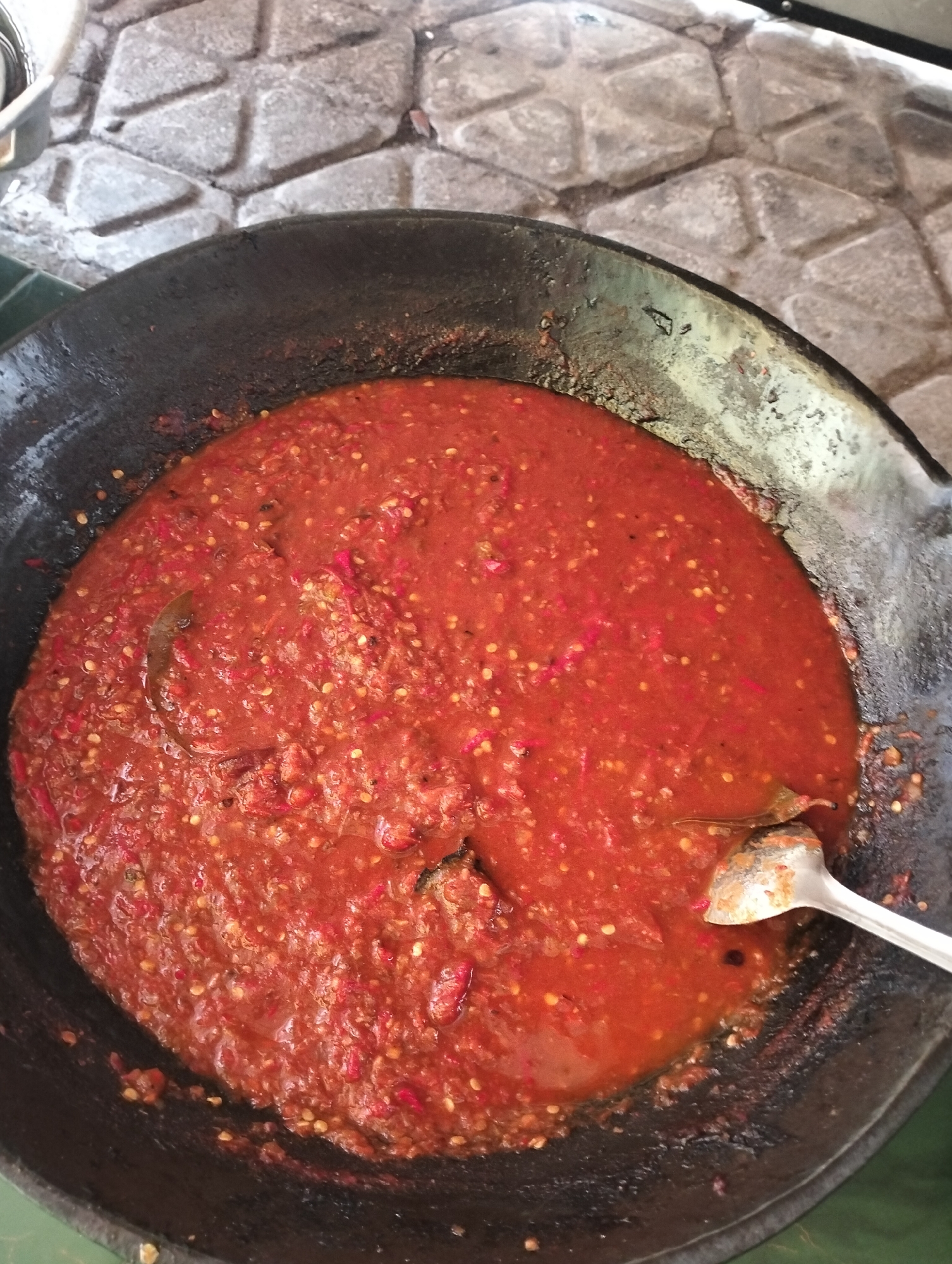 An overhead, close-up shot shows a large, dark, cast-iron wok filled with a thick, red chili paste. The paste is chunky and appears to be made from pureed chilies, with visible seeds and small pieces of chili flesh. A silver spoon rests on the edge of the wok, partially submerged in the chili paste. The wok is positioned on a textured, gray surface that resembles paving stones. The scene is lit with natural light, casting subtle shadows. The image appears to have been taken in an outdoor setting, possibly in Purwokerto, Indonesia, given the style of the wok and the chili paste, which is a common ingredient in Indonesian cuisine.