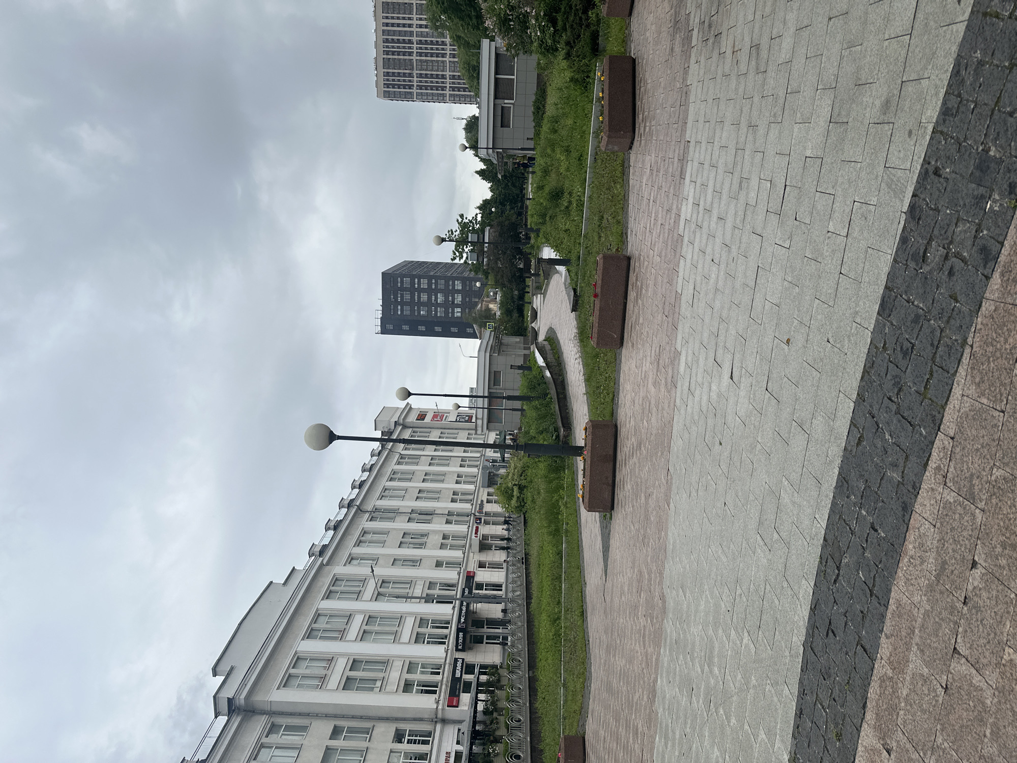 The image captures an urban street scene in Khimki, Russia, on an overcast day. The perspective looks along a paved walkway towards a complex of buildings.
In the foreground and to the right, a wide pedestrian path features distinct sections of light gray rectangular paving tiles, a curving band of dark gray cobblestones, and a section of reddish-brown square pavers. Rectangular planters, some containing small plants, line the walkway, accompanied by street lamps with spherical globes.
To the left, a large, multi-story white building with numerous windows dominates the midground. Several commercial signs are visible on its facade, including "Кухни" (Kitchens), "ШКОЛА" (School), "ЧИСТЫЙ БЕРЕГ" (Clean Shore/Bank), and a partially visible "ИМПЕРИЯ" (Empire), alongside a red logo resembling "TELE2". Bicycles are parked near the building.
Beyond the white building, a strip of green grass and trees separates it from other structures. In the middle distance, a tall, dark gray rectangular building stands out, and further back, another multi-story, lighter-colored high-rise is partially visible against the cloudy sky. The sky is uniformly gray and overcast, suggesting diffused daylight. No explicit human activity is taking place, but the urban infrastructure implies a functional city environment.