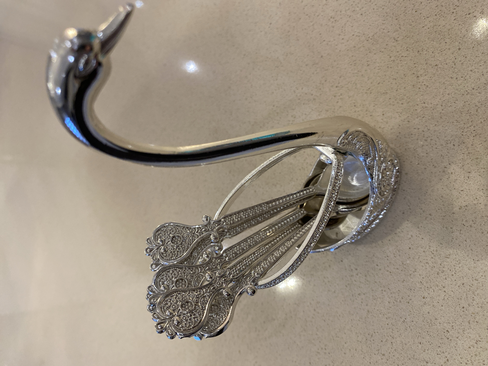 The image shows a silver-colored, decorative wall hook, possibly made of metal, mounted on a light-colored, speckled countertop or wall surface. The hook's design includes a curved, swan-like head and an ornate base containing several small, intricately detailed spoons. No people, actions, or interactions are visible in the frame. The location, Alanya, Turkey, cannot be verified from the visual information provided.