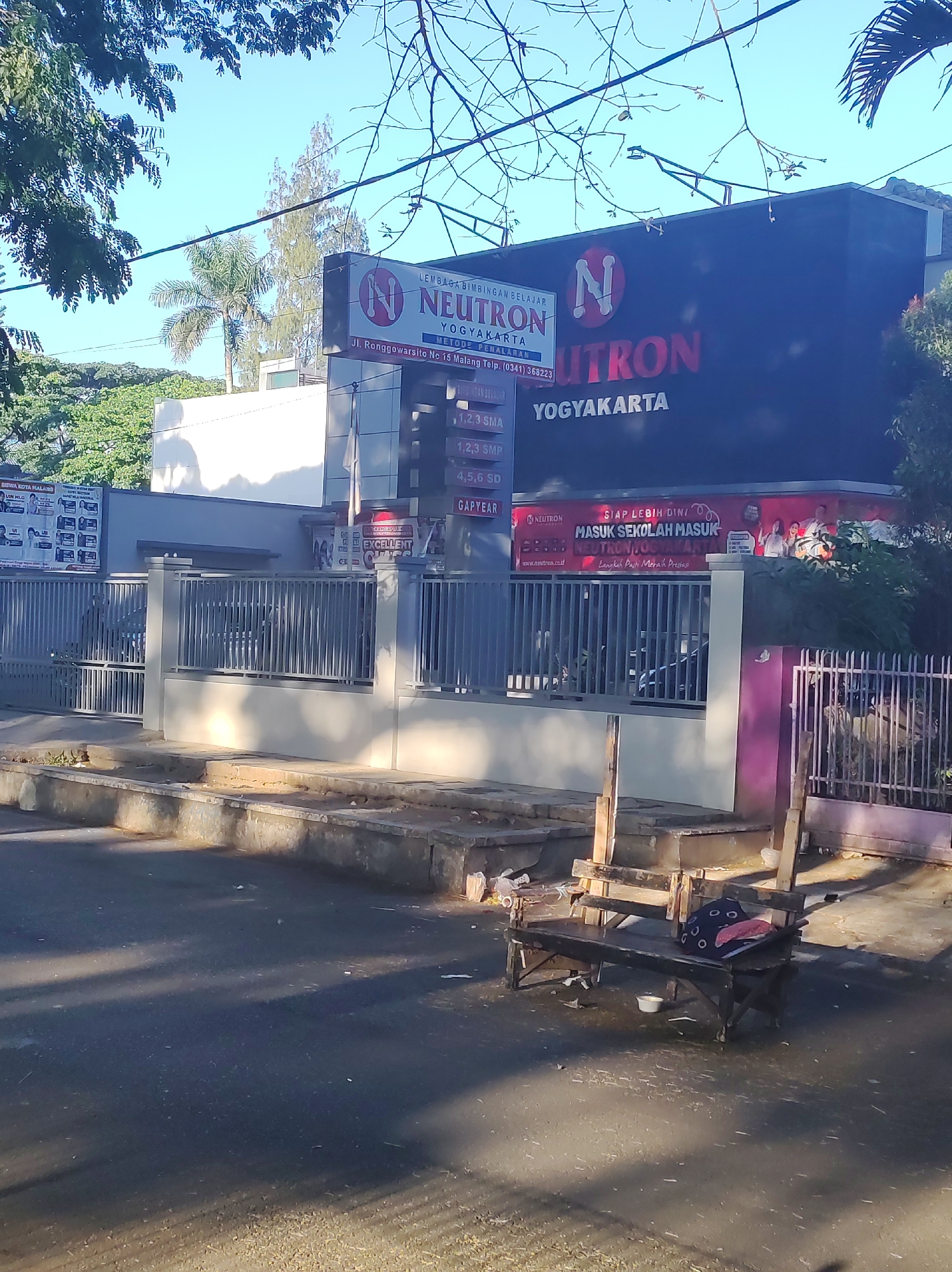 The image shows the exterior of a building in Malang, Indonesia, that houses a learning center called "Neutron Yogyakarta." The building has a dark facade with large, illuminated lettering that reads "NEUTRON YOGYAKARTA." A smaller sign above the entrance displays the learning center's name and address: "Jl. Ronggowarsito No. 15 Malang Telp. (0341) 368223." Below this, a list of educational programs is visible, including "1,2,3 SD," "1,2,3 SMP," and "4,5,6 SD," as well as "GAP YEAR." A banner on the right side of the building features the slogan "SIAP LEBIH DINI MASUK SEKOLAH MASUK NEUTRON YOGYAKARTA." The entrance is bordered by a metal fence. In the foreground, a street with a curb is visible, and a simple wooden bench with some items on it sits on the sidewalk. The scene is illuminated by natural sunlight, casting shadows on the street and sidewalk. Trees and some foliage are present in the background.
