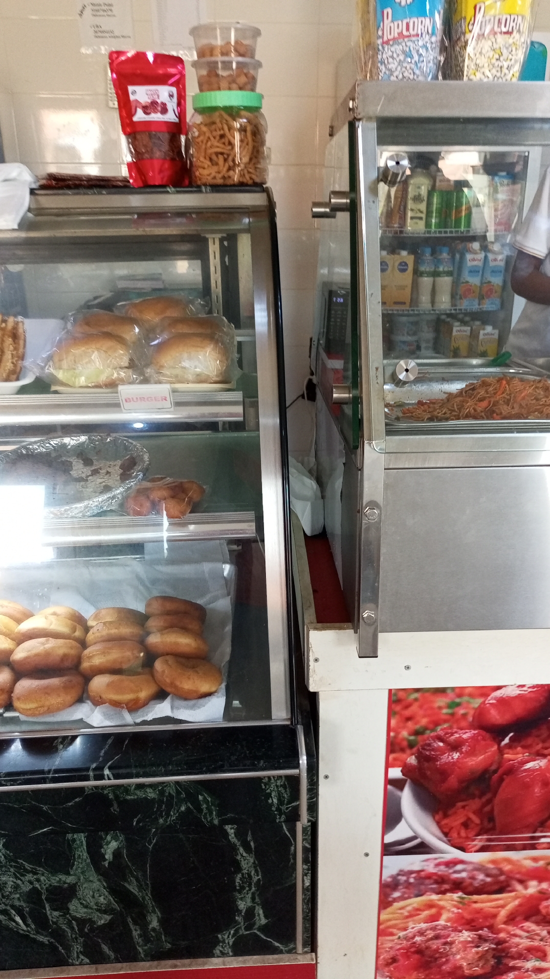 The image shows two refrigerated display cases side-by-side in what appears to be a food service establishment.
The case on the left is black with green marble pattern and contains:
- A top shelf with wrapped burger buns with a "BURGER" label.
- A middle shelf with a covered foil tray and some other fried items.
- A bottom shelf filled with fried dough items, possibly donuts.
On top of this case are a bag of red peppers, a jar filled with snack crackers, and smaller containers of food.
The case on the right is stainless steel and white. It has:
- A top shelf with drinks and boxes of popcorn.
- A middle shelf with an item that resembles noodles.
- A visible poster on the lower half of the unit with pictures of spaghetti and meatballs, as well as reddish-orange chicken on a bed of rice.
The image appears to be taken in Narayi, Nigeria, though there is no way to confirm this from the provided image details.