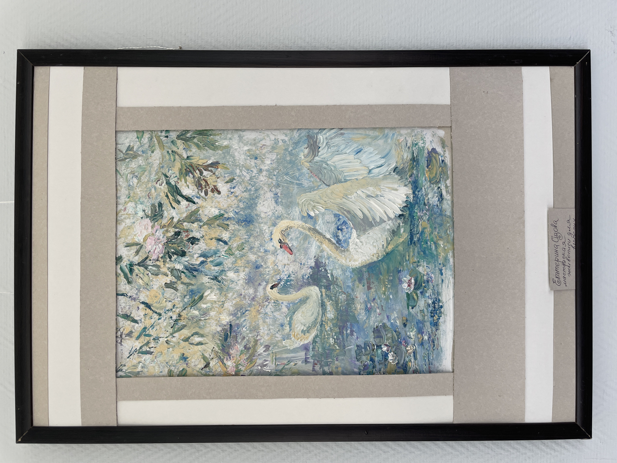 This is a framed painting of two swans in water with flowers and foliage. The painting uses thick brushstrokes, creating a textured appearance. One swan is larger and positioned in the upper right, with its wings outstretched as if in flight or landing. The second swan is smaller and positioned below and to the left of the first, appearing to be swimming or gliding. The water is depicted with shades of blue, green, and white, with hints of darker blues and purples that could represent shadows or depth. The surrounding vegetation is a mix of greens and earthy tones, with small touches of pink and white flowers scattered throughout. A small, light-colored label with handwritten text is attached to the right side of the matting. The text is in Cyrillic script and appears to be a name and possibly a title or description of the artwork. Based on the visible characters, it likely reads "Екатерина Пукова" and below that "мастерская где живут бабочки" which translates to "Ekaterina Pukova" and "workshop where butterflies live". The overall scene evokes a tranquil and natural setting, possibly a pond or lake during the daytime, with a focus on the grace and beauty of the swans. The framing consists of a black outer frame with a layered matting of grey and white paper strips.