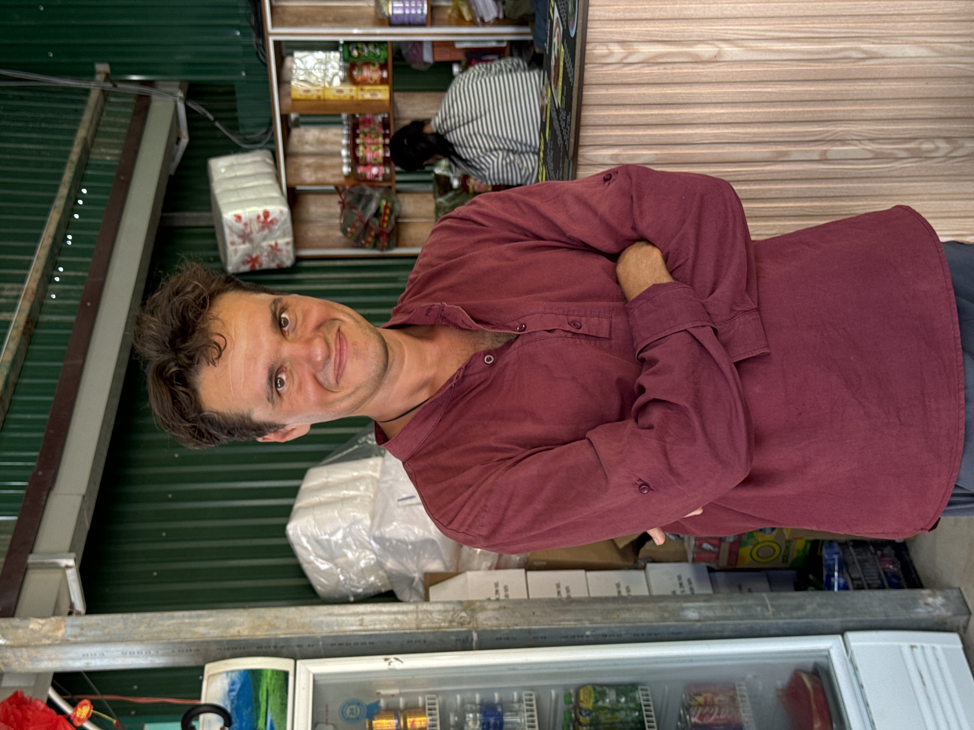 The image depicts a man in Phan Son, Vietnam, likely standing inside a small convenience store. He is fair-skinned, with dark hair and eyes, wearing a long-sleeved, maroon shirt and is standing with his arms folded. Behind him, there is a green, corrugated metal wall. In the background, there is another individual with dark hair wearing a striped shirt, partially obscured by shelving filled with various packaged goods. Below the man is a refrigerated display case stocked with food and beverages.