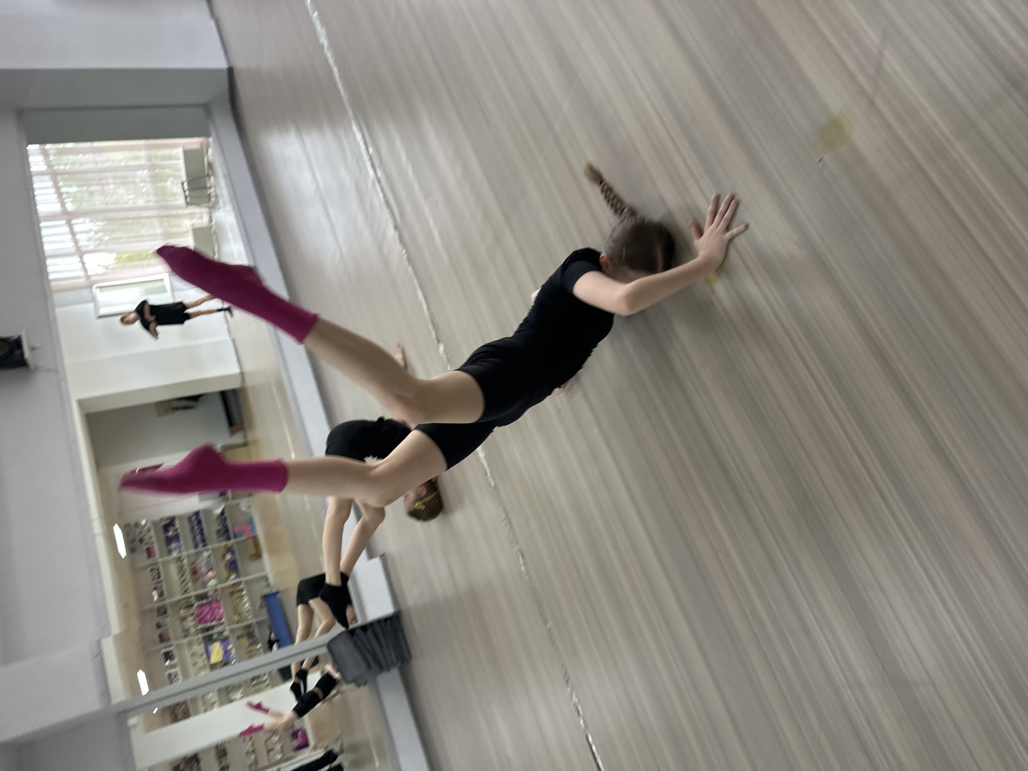 The image shows a gymnastics training session in a mirrored studio. A young gymnast wearing a black leotard and pink socks is performing a handstand split against a wall. Another gymnast is visible in the background, near a mirror. Shelves filled with trophies and shoes line the wall behind the gymnasts. The studio has a light-colored floor with a textured, wood-like pattern. Based on the context provided, the location is likely in Votkinsk, Russia.