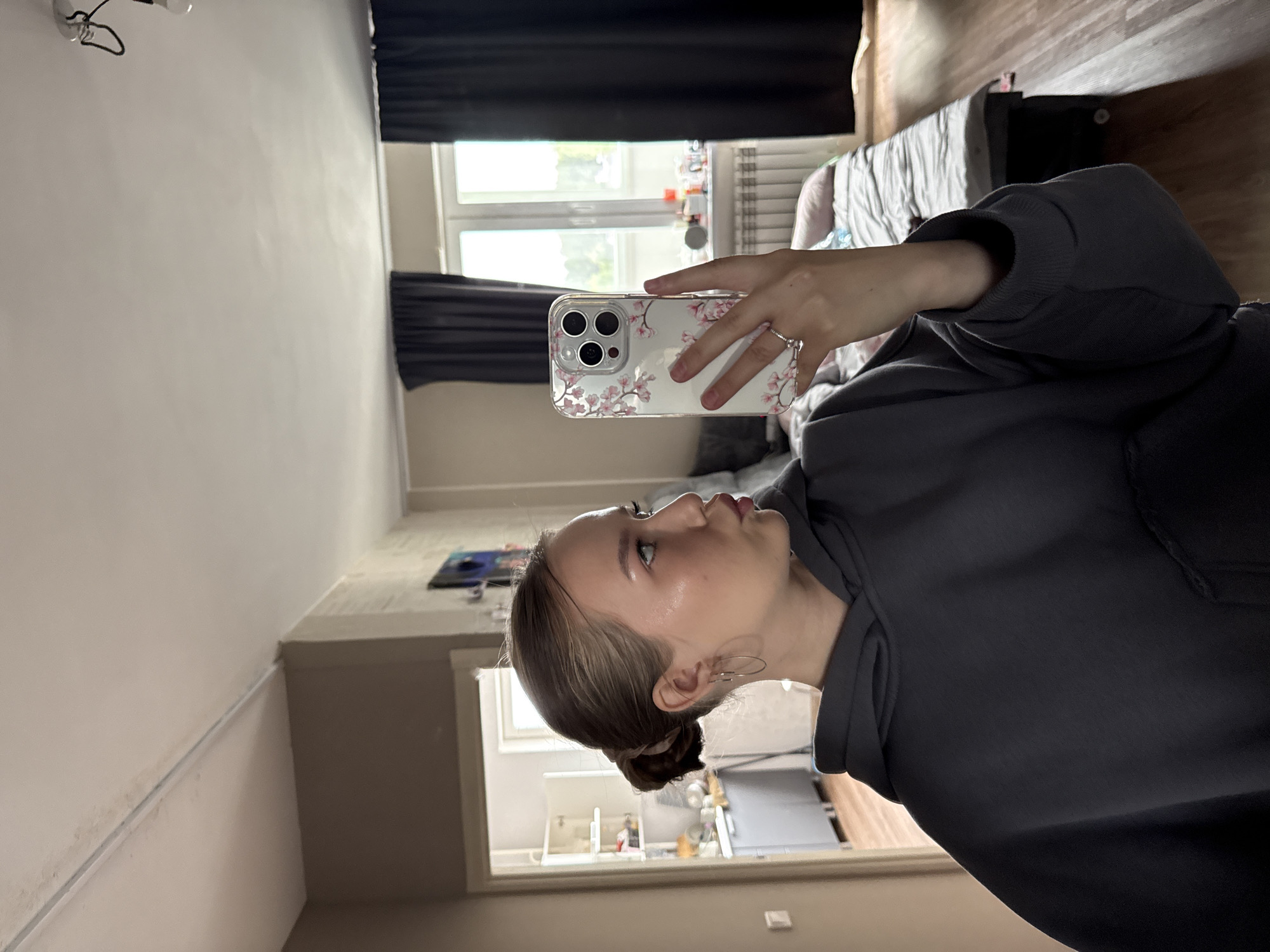 A young woman is captured in a mirror selfie, seen in profile as she looks towards the smartphone she holds in her right hand. Her hair is styled in a bun, and she wears a dark grey long-sleeved top, hoop earrings, and a ring on her right ring finger.
The smartphone she uses has a clear case decorated with delicate pink floral patterns, reminiscent of cherry blossoms, and features a triple-lens camera system on its rear. The reflection in the phone's screen shows a window with dark blinds or curtains, indicating a daytime setting with natural light.
The background reveals an indoor residential room, likely a bedroom or living area in Chaikovsky, Russia. It features light-colored walls, a window with dark coverings, and a glimpse of a bed with light bedding. A light fixture is visible on the ceiling, and various indistinct objects populate surfaces within the room. The overall ambiance is casual and domestic. No visible text is discernible in the image.
