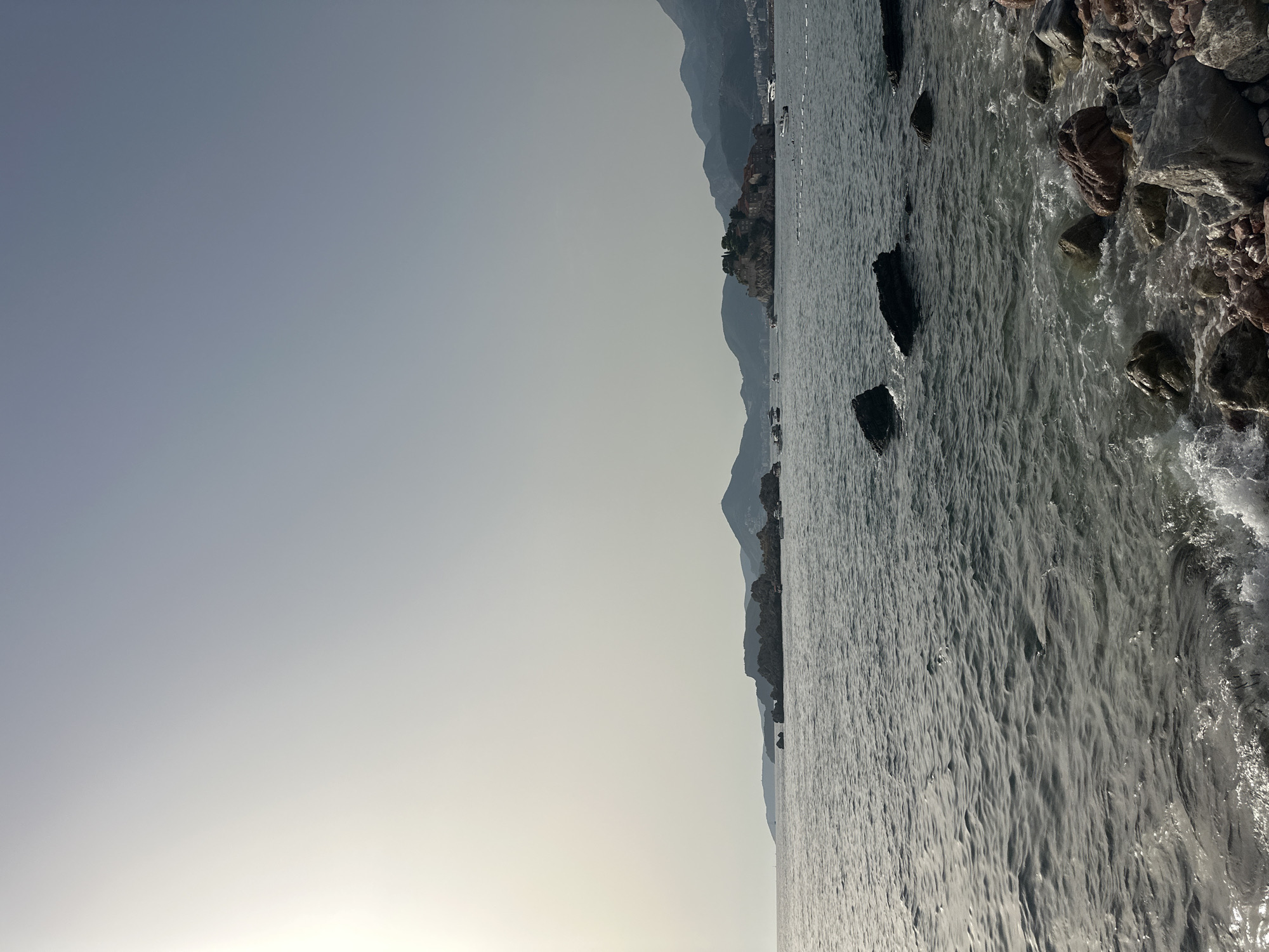 A wide shot captures a tranquil seascape near Sveti Stefan, Montenegro, under a hazy, bright sky. The foreground features a rocky shoreline with various-sized stones, some of which are partially submerged in the gently lapping water. The water itself is a deep grey-blue, with subtle ripples reflecting the overcast sky. Several dark, jagged rocks break the surface of the water further out. In the mid-ground, a cluster of rocky islets, one of which is crowned with the red-tiled roofs and walls of historic buildings, forms the iconic Sveti Stefan. Boats of various sizes are visible in the water around the islets and along the distant coastline. The background is dominated by the silhouettes of rolling, mountainous terrain that fades into the hazy horizon. The overall atmosphere is serene and somewhat muted due to the diffused sunlight.