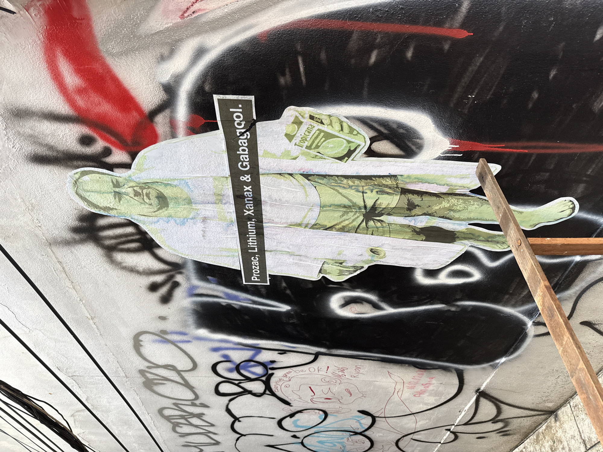 The image shows a close-up of a graffiti-covered wall, likely in an urban setting. The wall is covered with various layers of graffiti, including large black and white abstract shapes, red streaks, and other colorful tags. A sticker or print has been pasted onto the wall, featuring an image of a person lying down, wearing what appears to be a lab coat and shorts, with a Tropicana juice box near their head. A black strip with white text reads "Prozac, Lithium, Xanax & Gabapentol." Below this image, there is more graffiti, including a drawing of a face with the text "everything gonna be OK!" and a name that appears to be "Ryan." A wooden plank is leaning against the wall on the right side of the image. The photograph appears to be taken at an upward angle, looking at the underside of a structure, possibly a bridge or overpass. While the image hints at an urban setting and mentions Thailand, there is not sufficient detail to positively identify the location within Bangkok, Thailand.