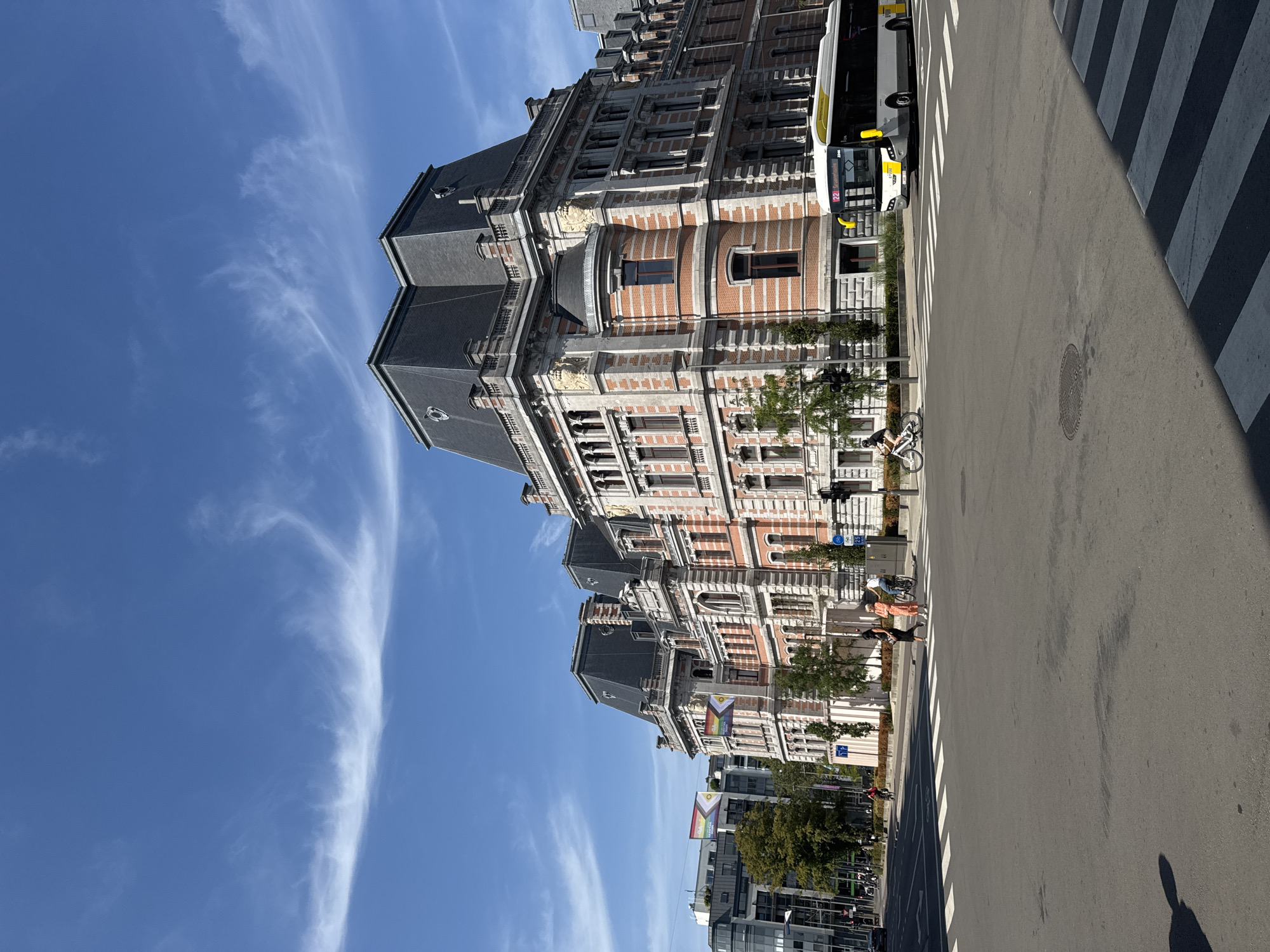 The image depicts a street scene in Antwerp, Belgium. A large, ornate, historic building with a dark mansard roof dominates the right side of the frame. The building features red brickwork with grey stone accents and numerous windows. In front of the building, a street intersection is visible with a crosswalk. A modern white and yellow bus is turning at the intersection. Several people are present: two women are walking across the crosswalk, and two individuals are riding bicycles on the street and sidewalk. In the background, other modern buildings and trees can be seen, along with a clear blue sky with scattered wispy clouds. Two flags with rainbow stripes are visible on a building in the background, suggesting a Pride event or celebration.